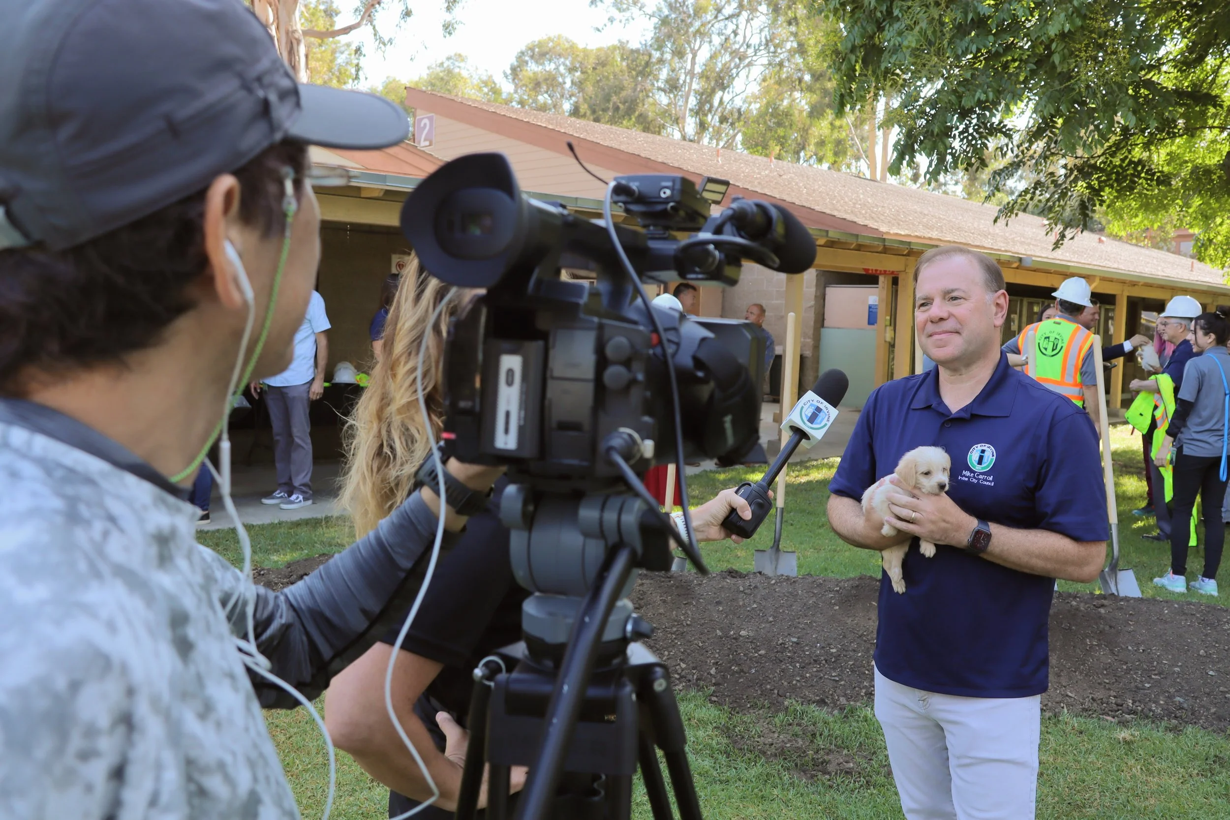 A man holding a puppy is being interviewed outdoors by a reporter with a video camera. People wearing construction helmets and reflective vests are visible in the background near a building under construction, with trees and a clear sky above.