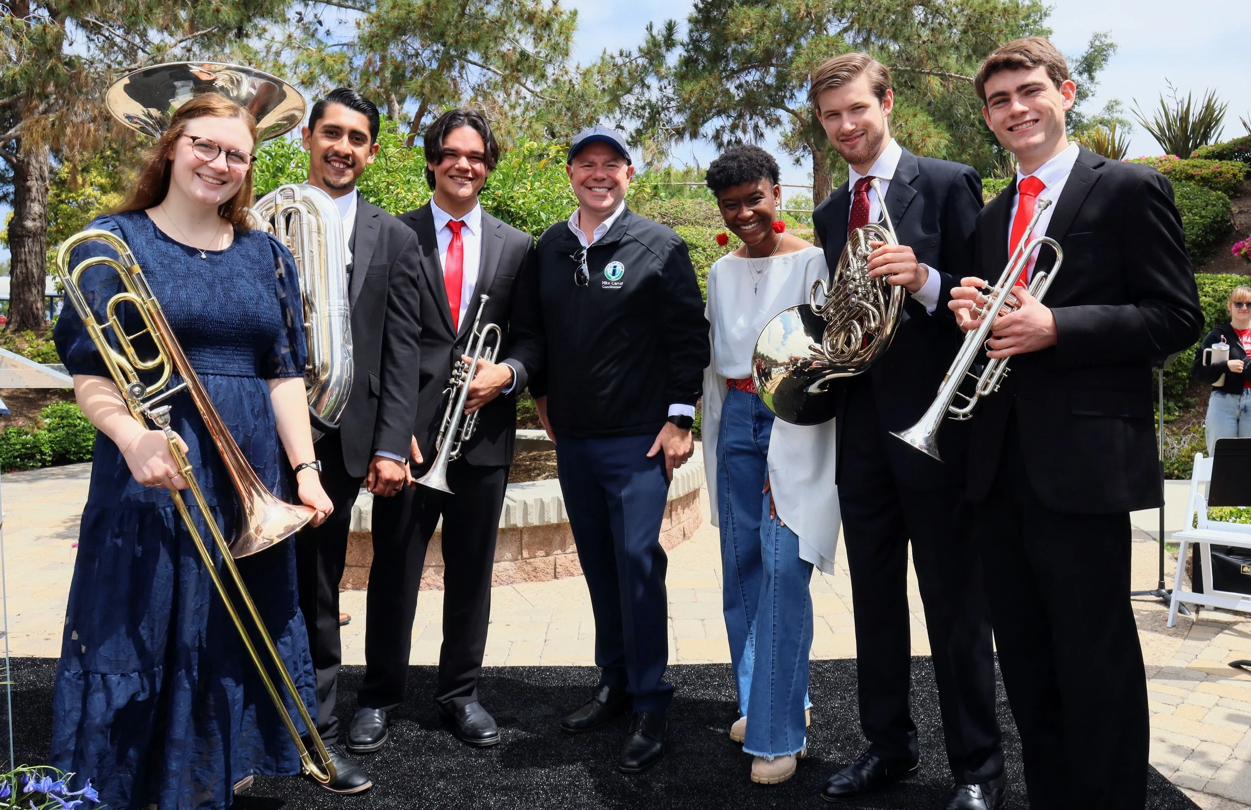 A group of seven musicians, six men and one woman, outdoors holding brass instruments, smiling at the camera with greenery and trees in the background.