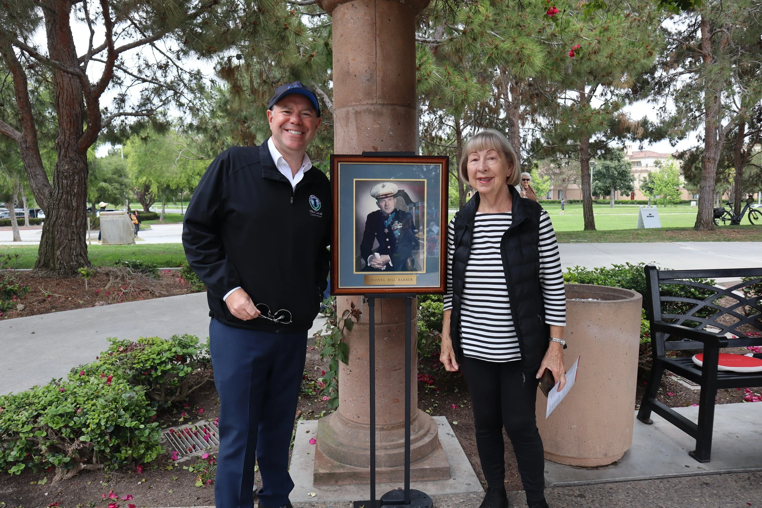 A man and a woman standing outdoors near a monument with a framed photograph of a military person labeled Colonel Bill Barber.