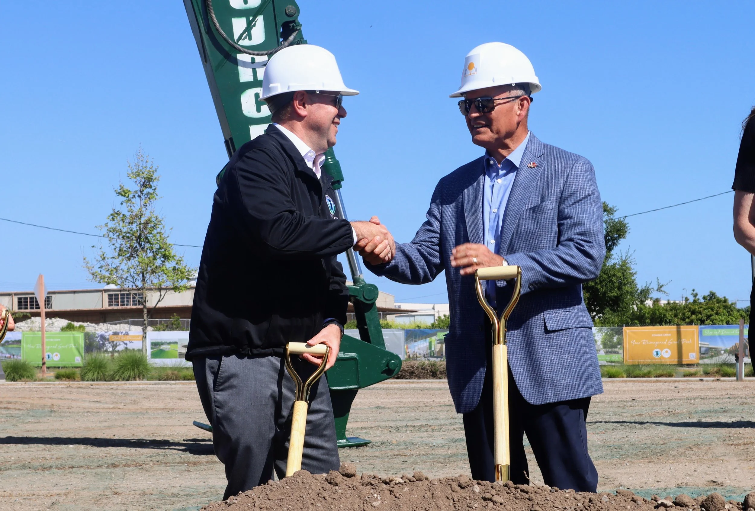 Two men wearing hard hats shake hands at a groundbreaking ceremony, each holding a shovel, on a clear day with a construction site in the background.
