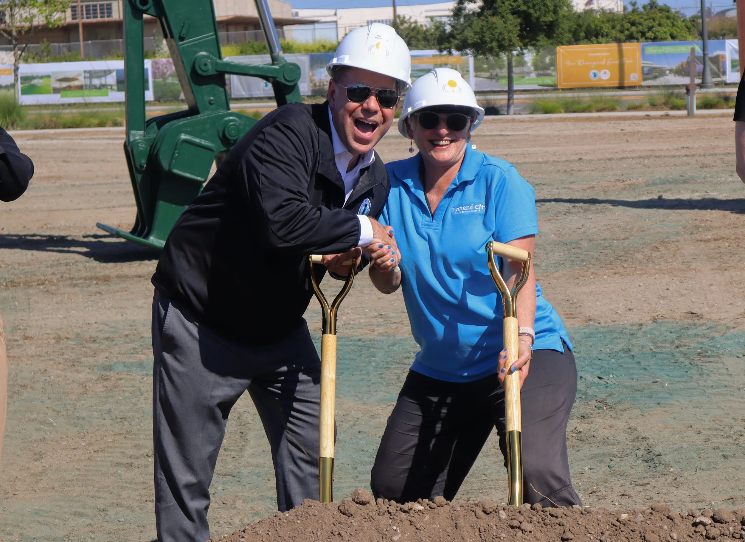 Two smiling people wearing hard hats and sunglasses, shaking hands, at a groundbreaking ceremony with shovels and dirt in an outdoor construction site.