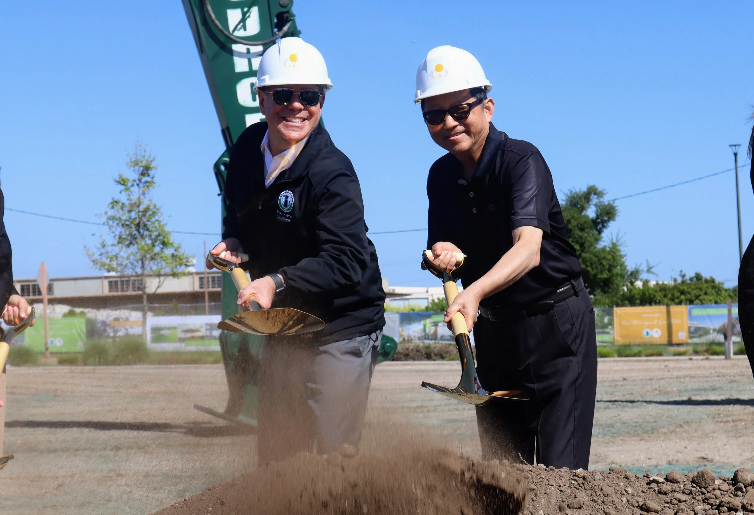 Two men wearing white hard hats and sunglasses, shoveling dirt at an outdoor construction site under a clear blue sky.