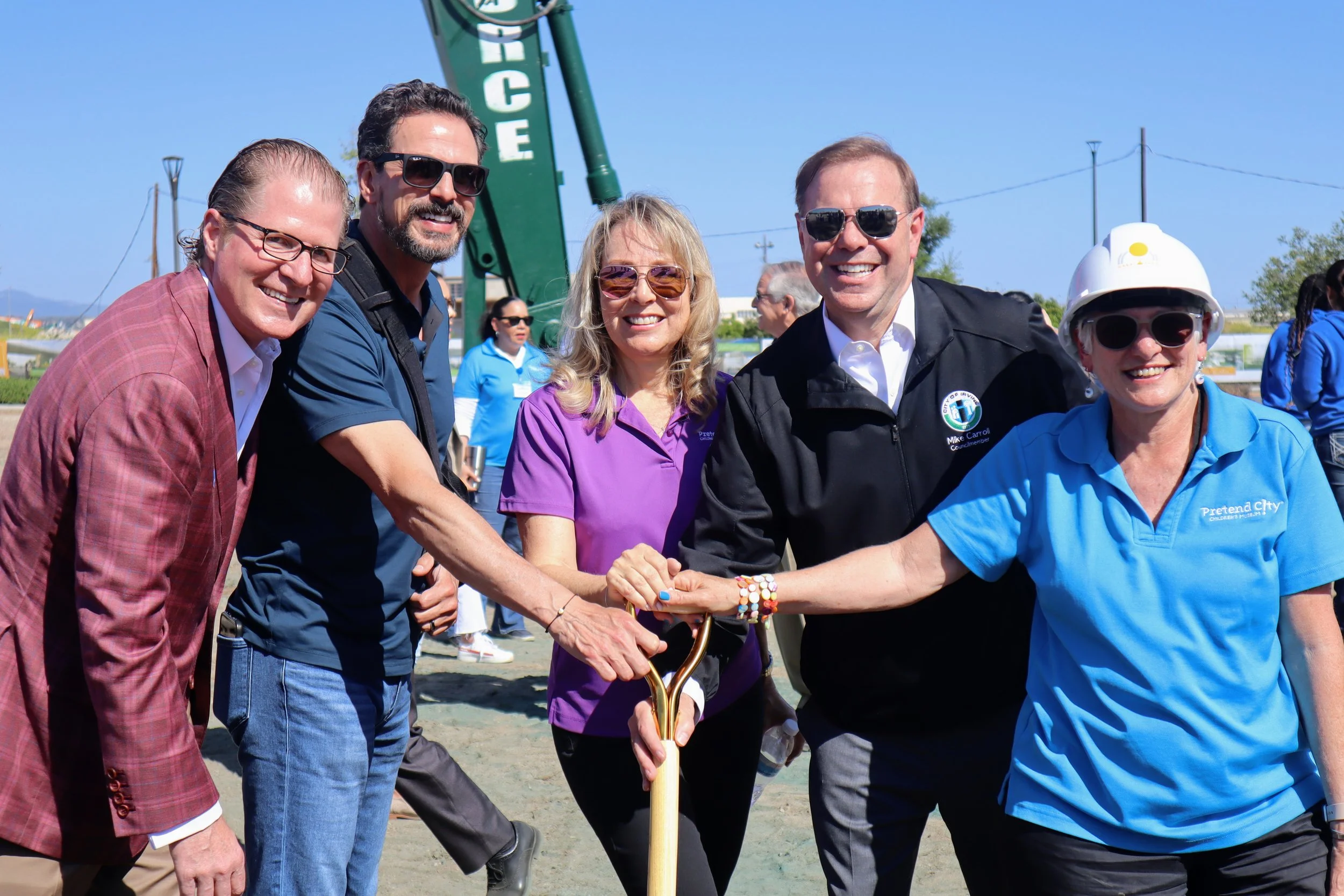 Group of five people at a groundbreaking ceremony, holding shovels together, smiling, outdoors on a sunny day.