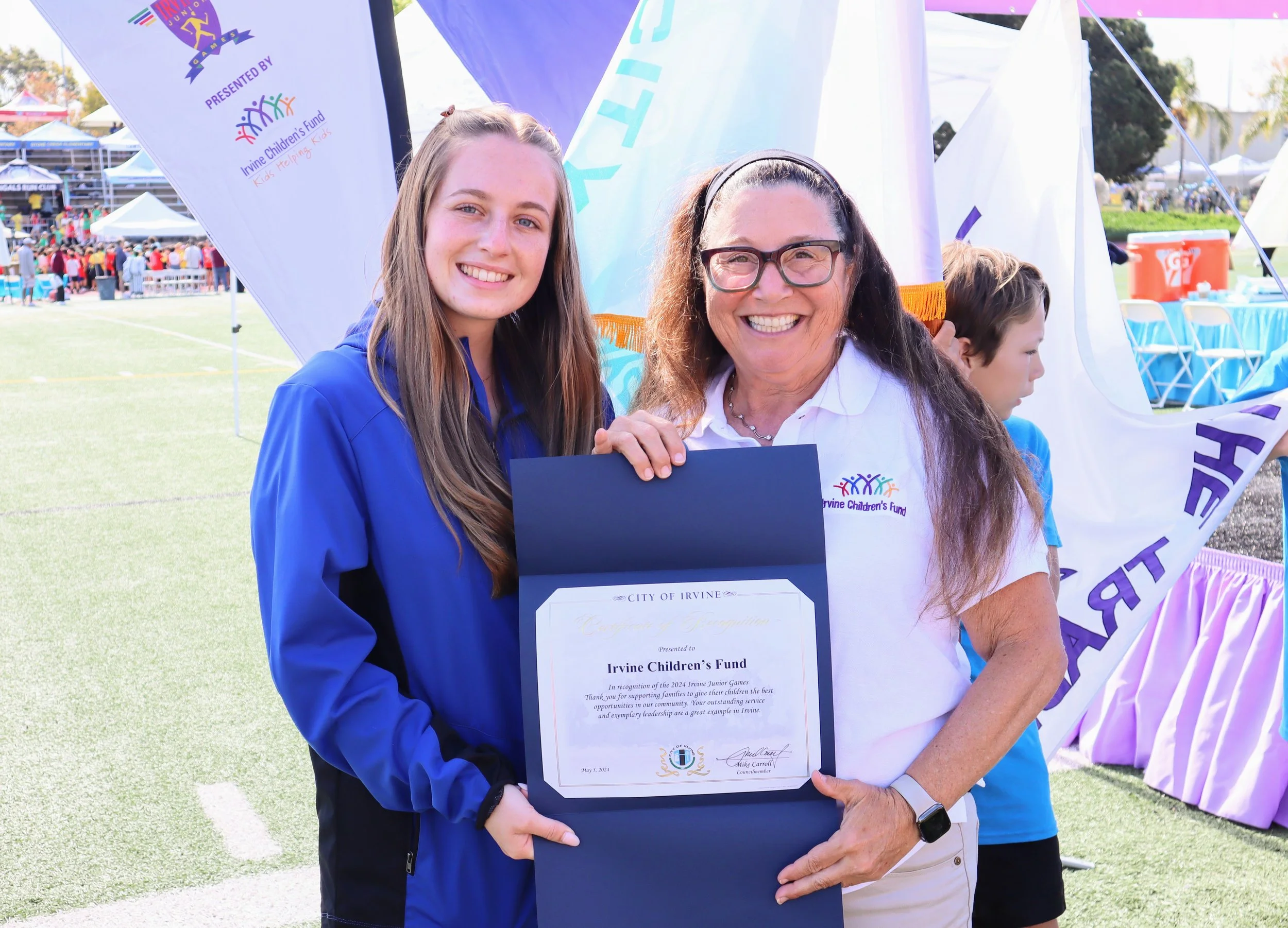 A young girl and an older woman are smiling and holding a certificate at an outdoor event. The girl has long, light brown hair and is wearing a blue jacket, while the woman has long curly hair, glasses, and is wearing a white shirt with a logo. There