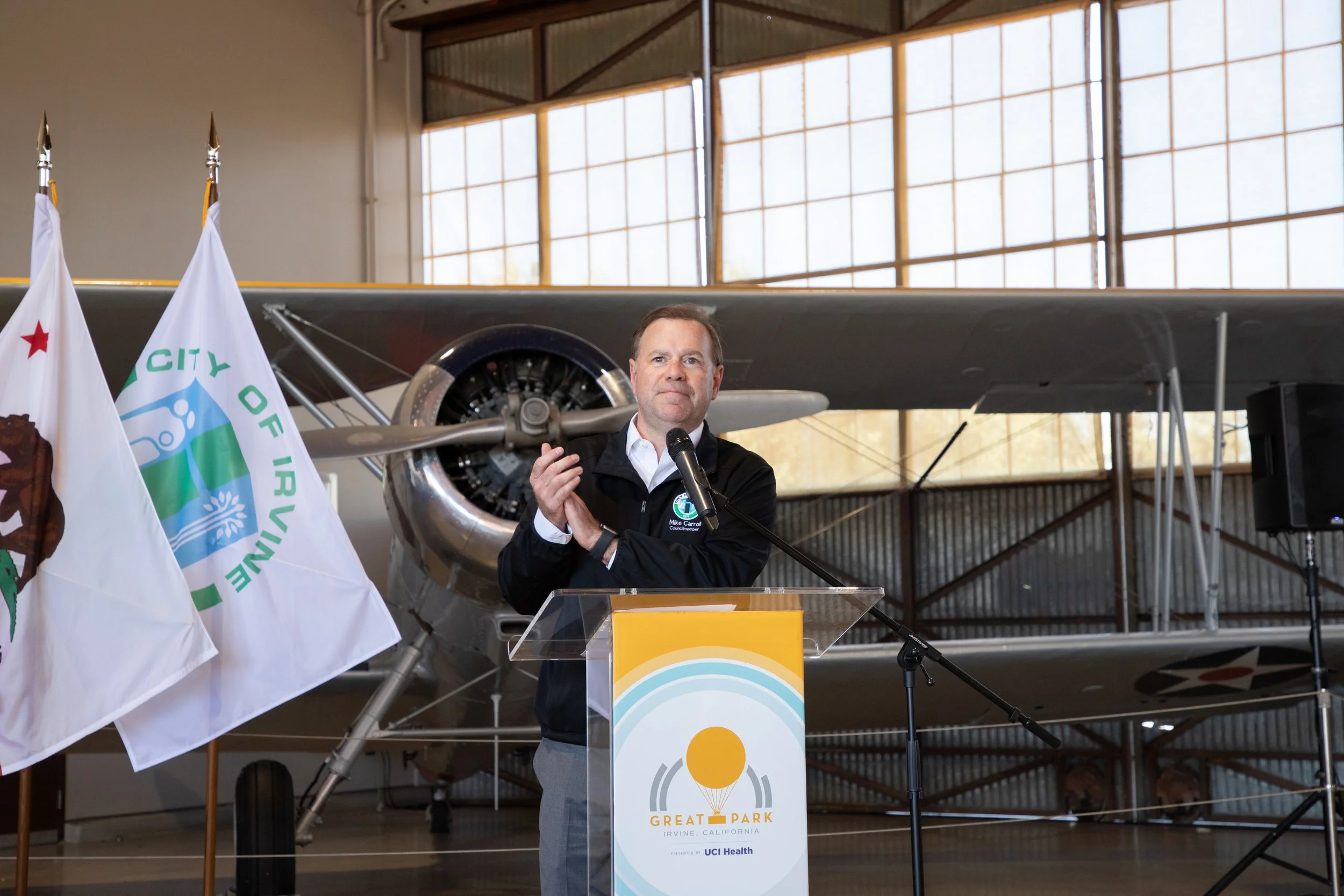 A man stands at a clear podium, speaking into a microphone, with flags and an airplane displayed behind him inside a hangar or aerospace museum.