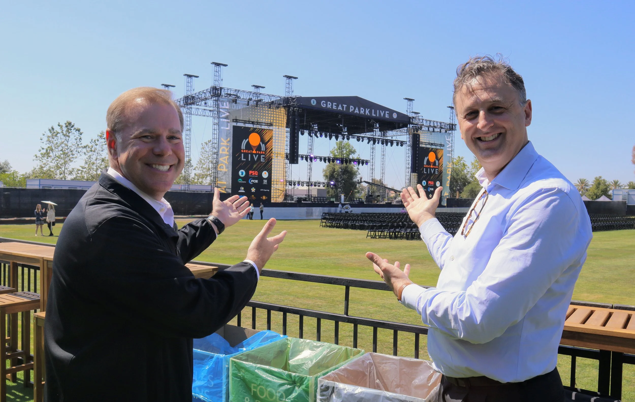 Two men standing outdoors in front of a stage at a park, smiling and gesturing towards the stage, with some trash bins in the foreground.