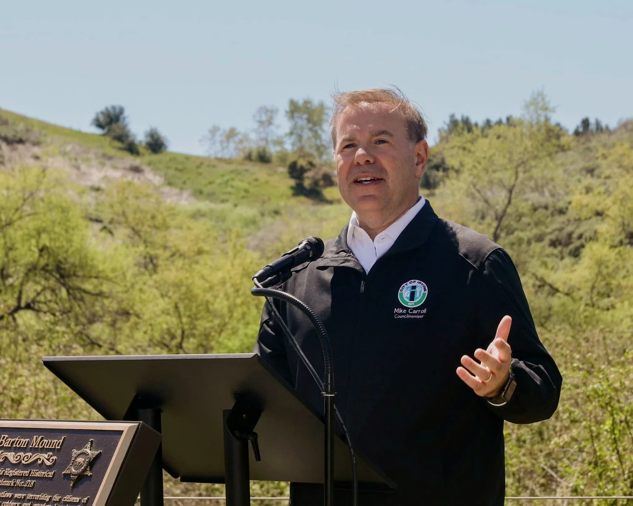 Man speaking at an outdoor event, standing behind a podium with a microphone, wearing a black jacket with a city seal and the name Mike Carroll, with green hills and trees in the background on a sunny day.
