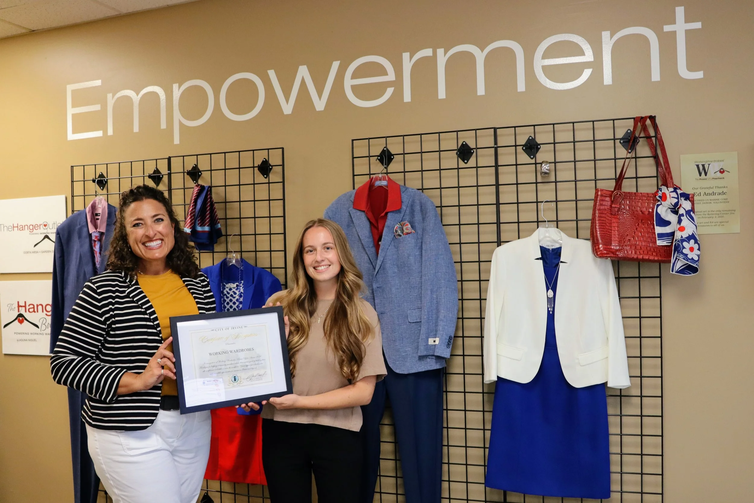 Two women smiling and holding a framed certificate in front of a display of professional clothing on grid panels, with the word 'Empowerment' on the wall behind them.