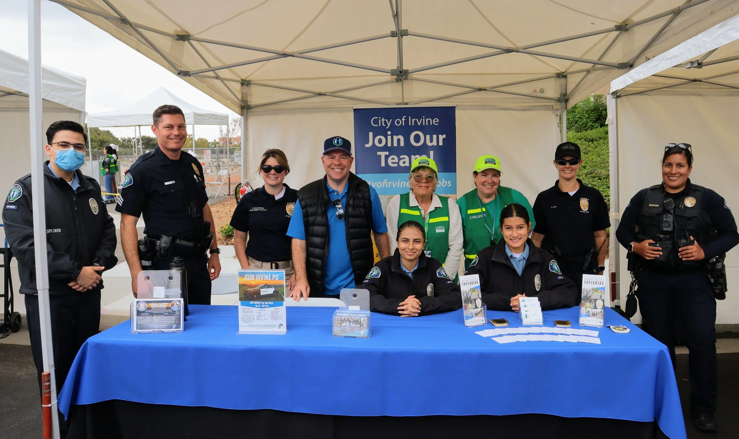 Group of police officers, city officials, and volunteers standing behind a booth with a blue tablecloth at a community event. There are flyers and pamphlets on the table, and a banner in the background reads "City of Irvine Join Our Team!".