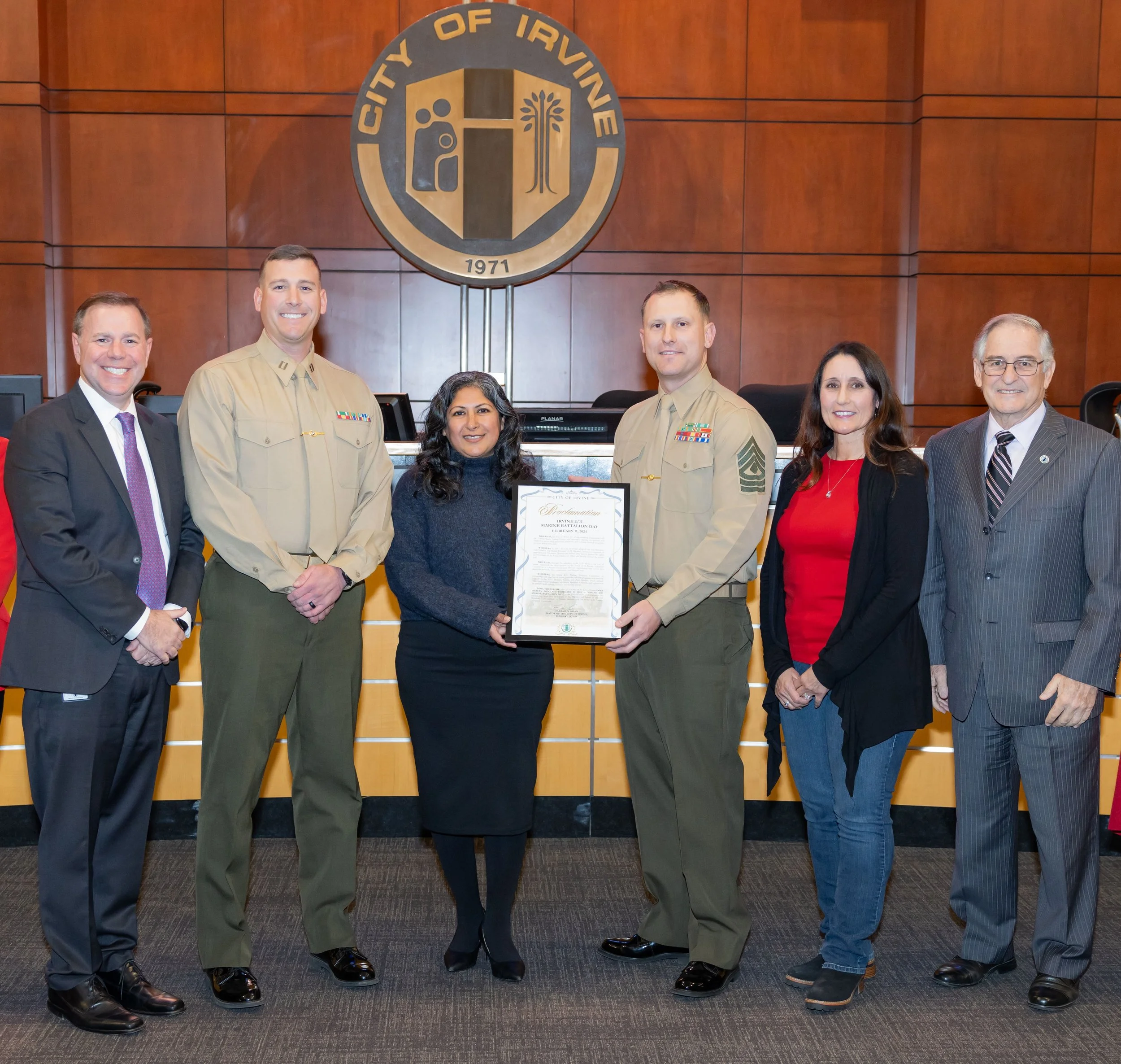 Group of seven people standing in a formal setting, with two in military uniform, holding a framed document, in front of a wooden wall with a large emblem reading 'City of Irvine'.