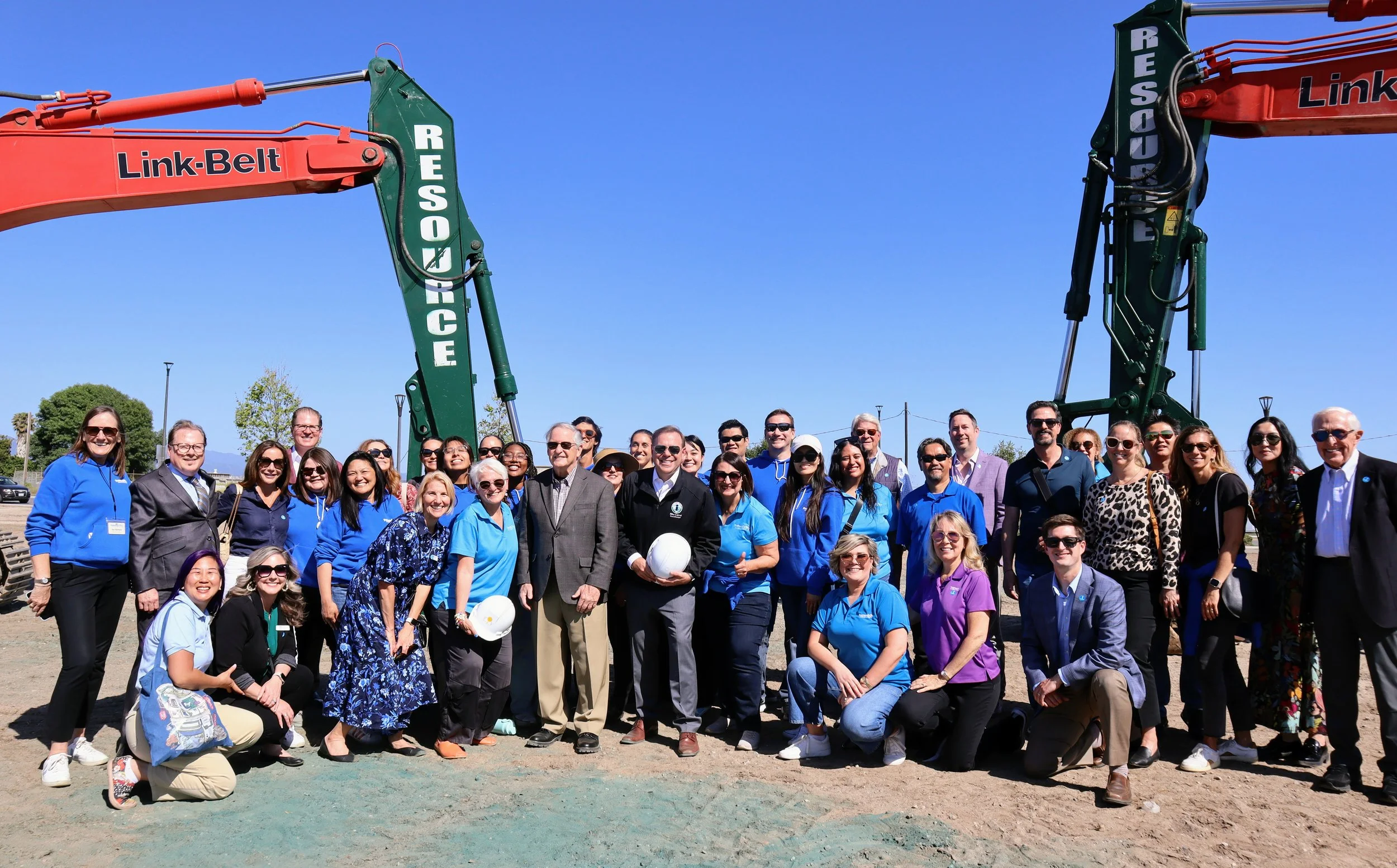 Group of diverse people gathered outdoors for a photo, standing in front of large excavators, with clear blue sky overhead.