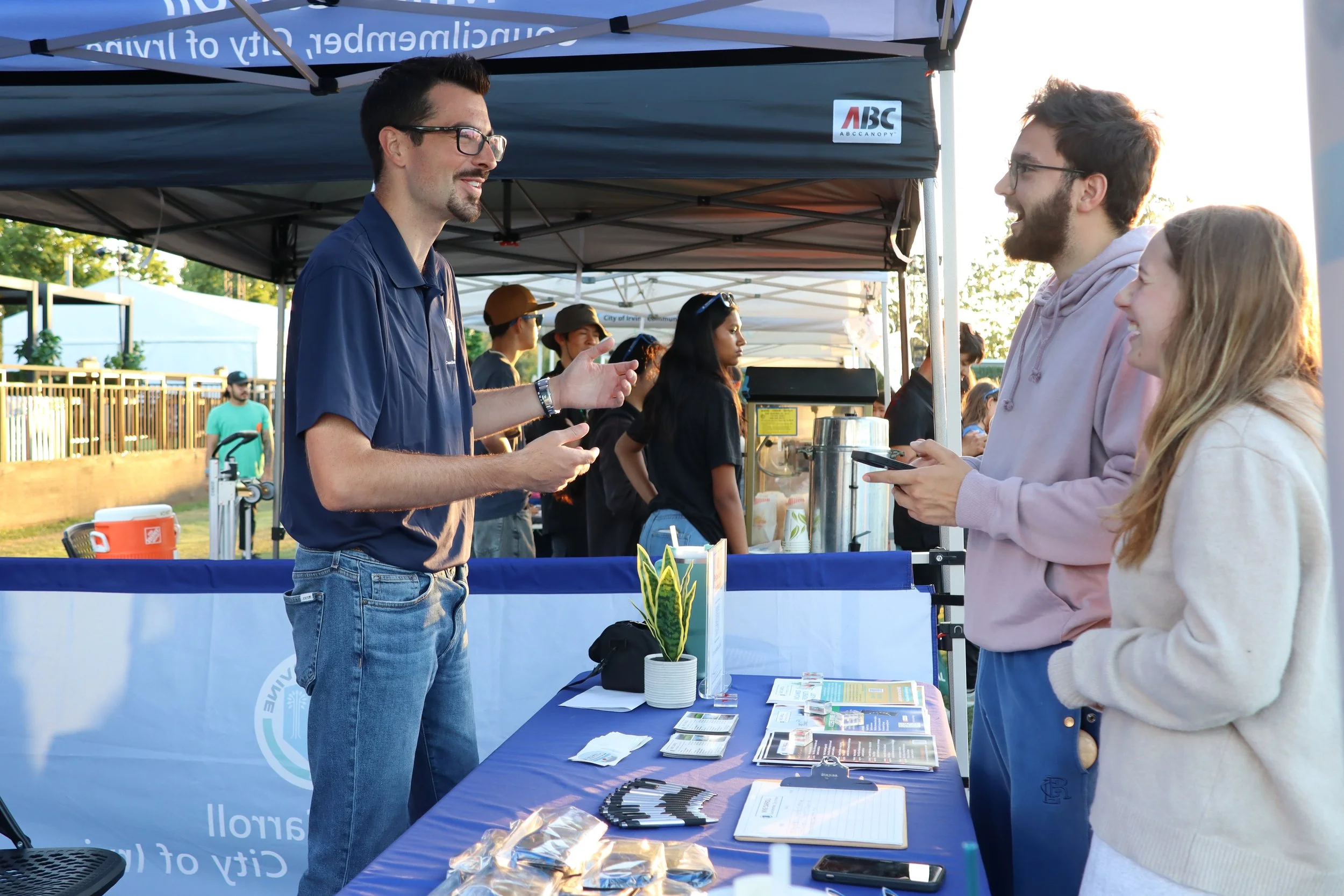 Three people engaged in conversation at an outdoor booth, with a man in a dark blue shirt and glasses talking to a man in a pink hoodie and a woman in a white coat, at a fair or event under a blue canopy.