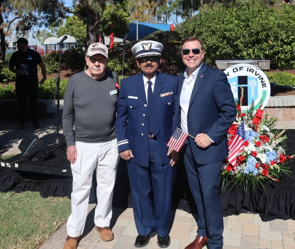Three men standing outdoors at an event with a floral arrangement and a sign that says 'City of Irvine' in the background. One man and one in a uniform are holding small American flags.