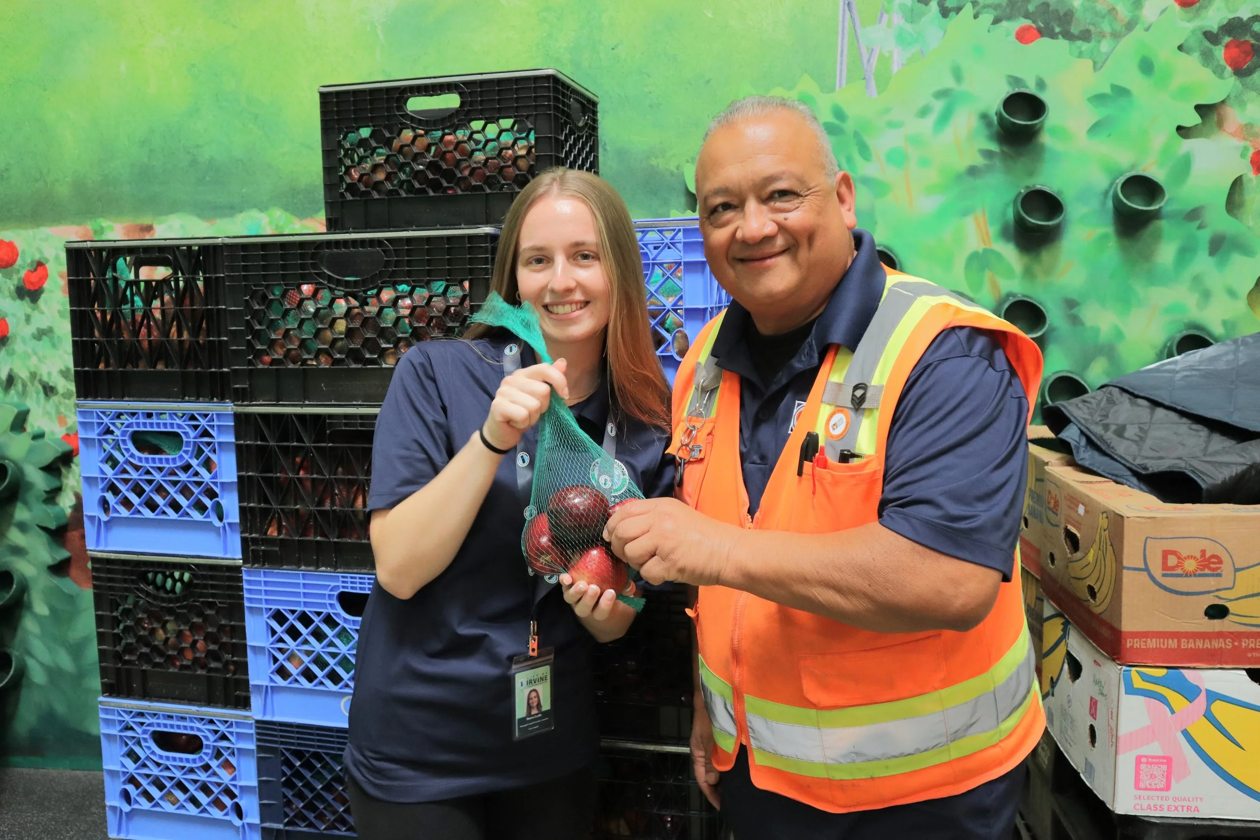 A woman and a man smiling at the camera while holding a bag of apples inside a warehouse or storage room with crates of apples and boxes of bananas in the background.