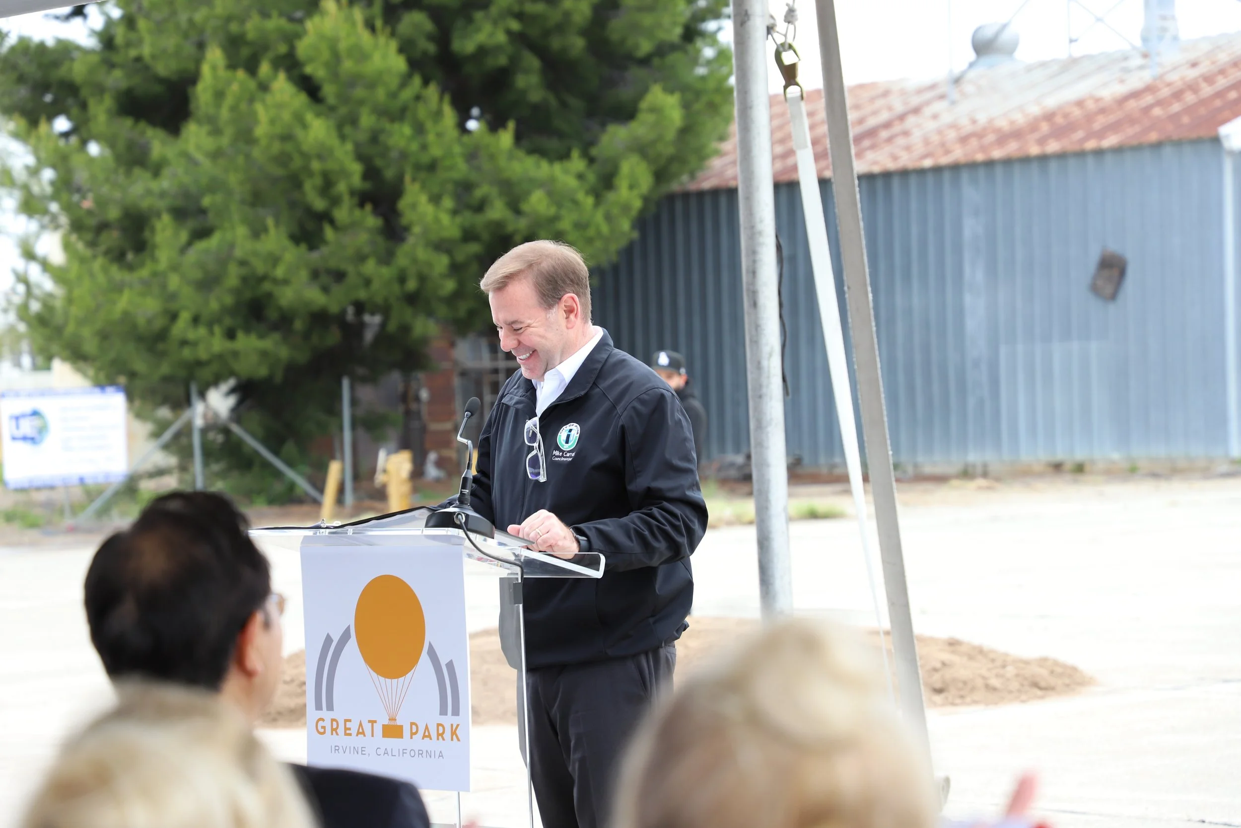 Mike Carroll for Great Park in Irvine, California, outdoors with trees, a fence, and industrial buildings in the background, and audience members visible in the foreground.