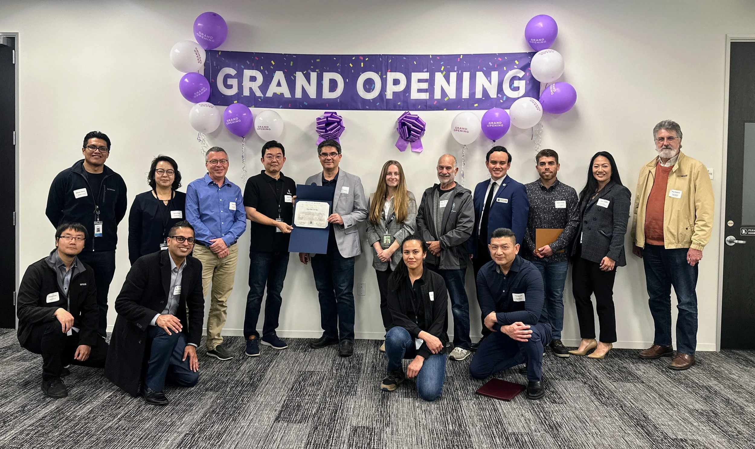 Group of people at a grand opening celebration, standing in front of a wall decorated with purple and white balloons and a banner that says 'GRAND OPENING'.