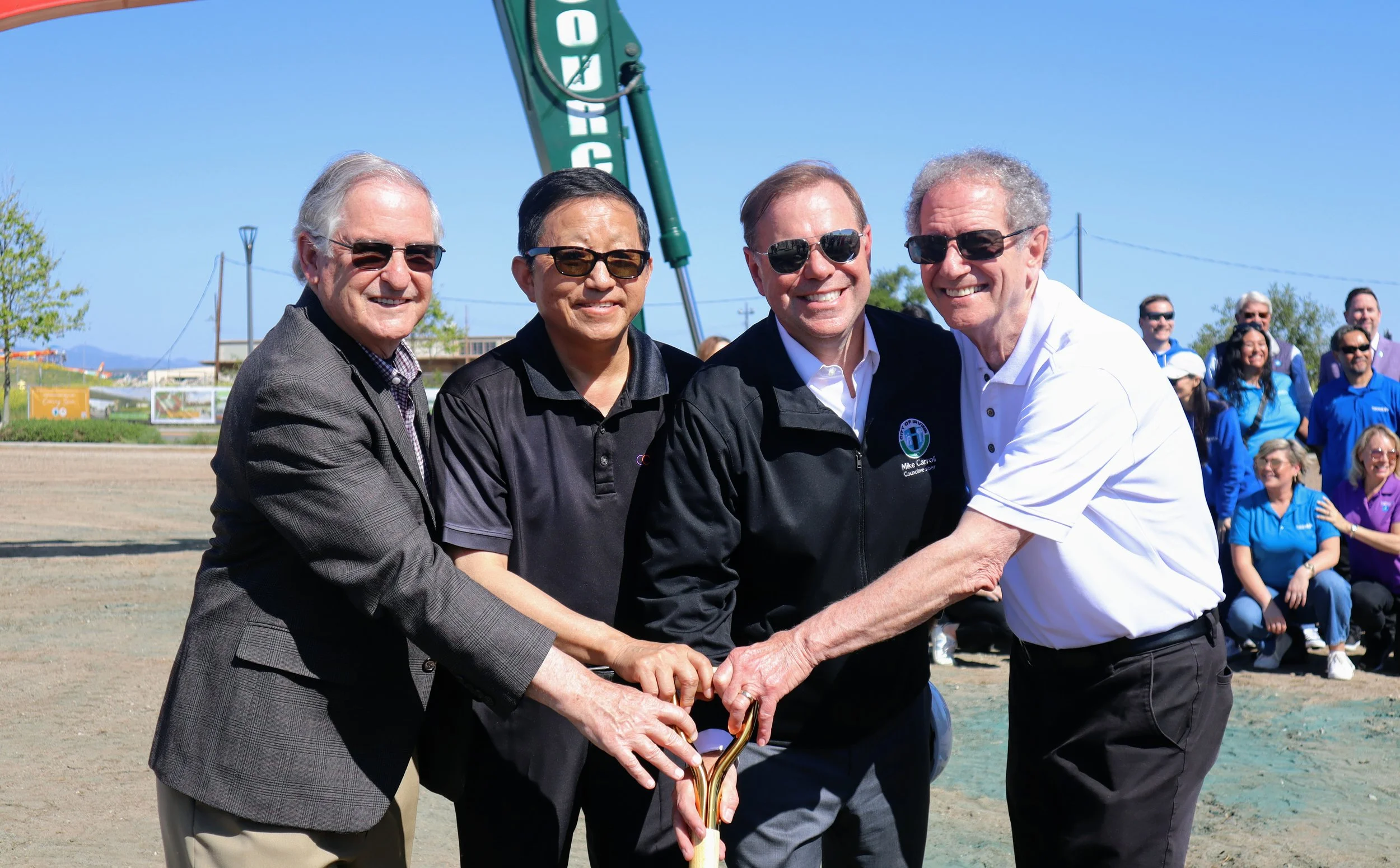 Four men in sunglasses and business casual clothing gather around a golden shovel during a groundbreaking ceremony, with a crowd of people in the background on a sunny day.