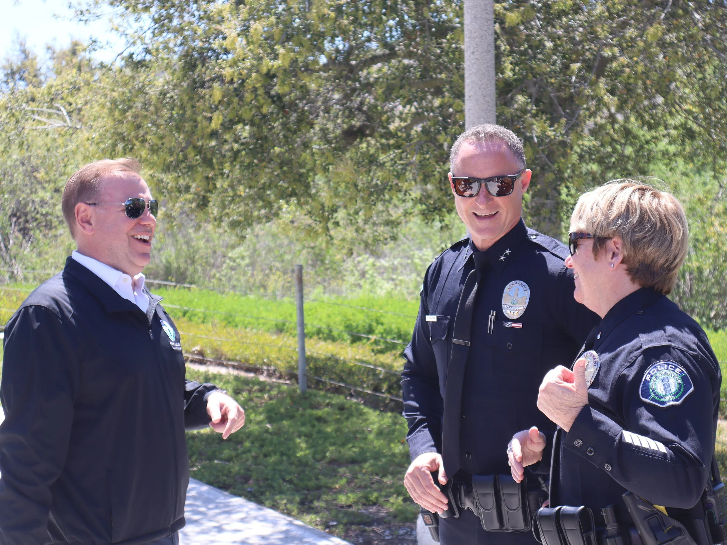 Three police officers, two men and one woman, engaging in a conversation outdoors during the daytime, all wearing dark police uniforms and sunglasses, with trees and greenery in the background.