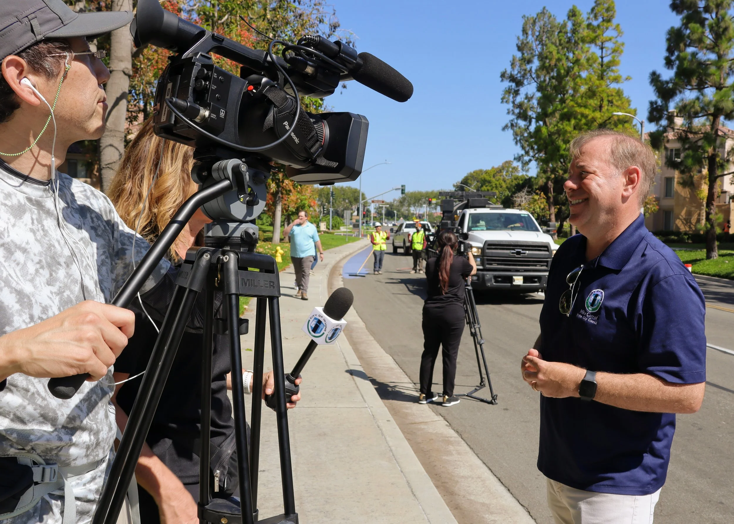 A man being interviewed outdoors by news crew, with cameras and crew members on the sidewalk, in front of a street with trees and traffic.