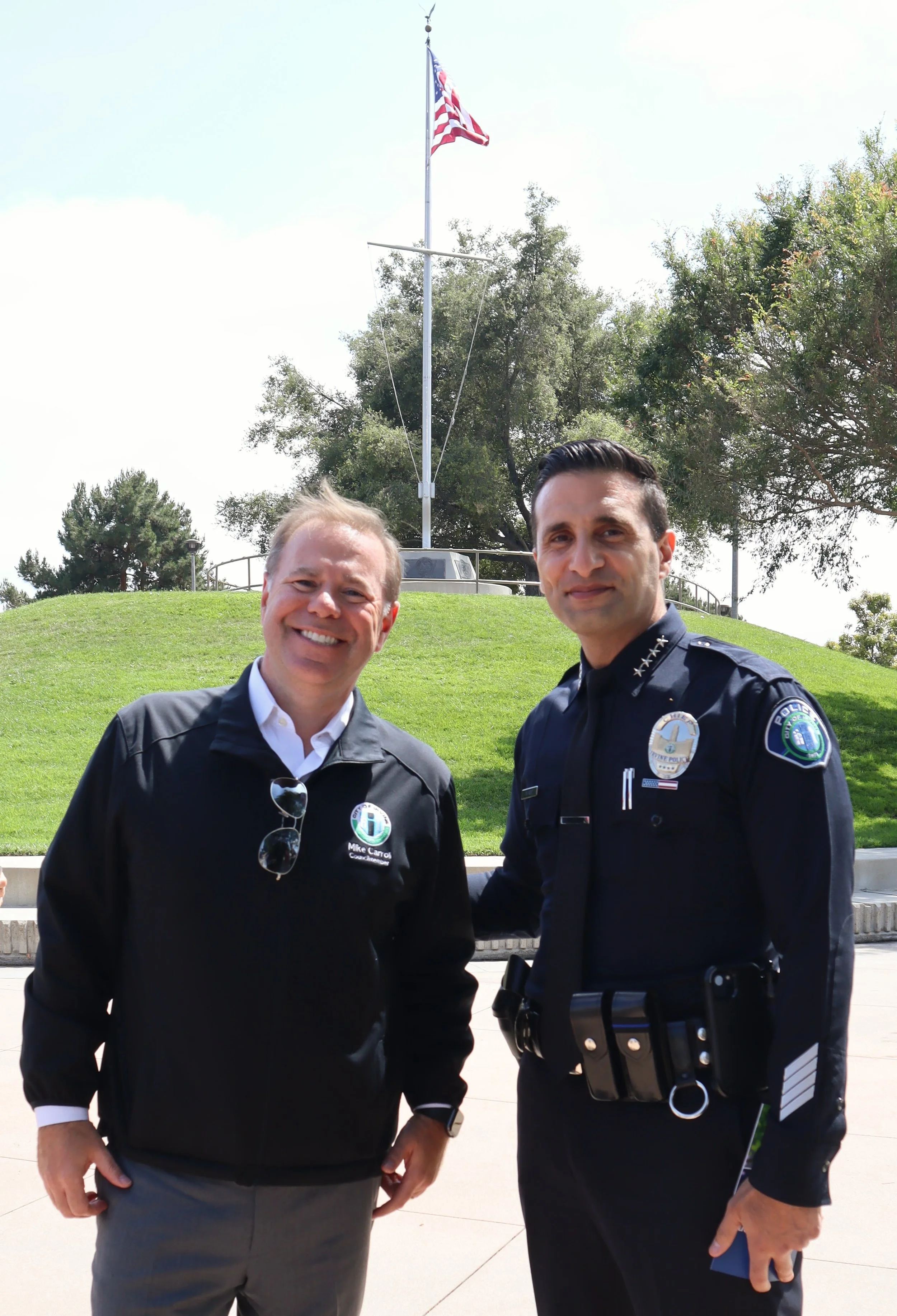 Two men standing outdoors near a grassy hill with an American flag on a pole. One man is dressed in a black jacket with a name tag, and the other is in a police uniform. They are smiling and standing close to each other.