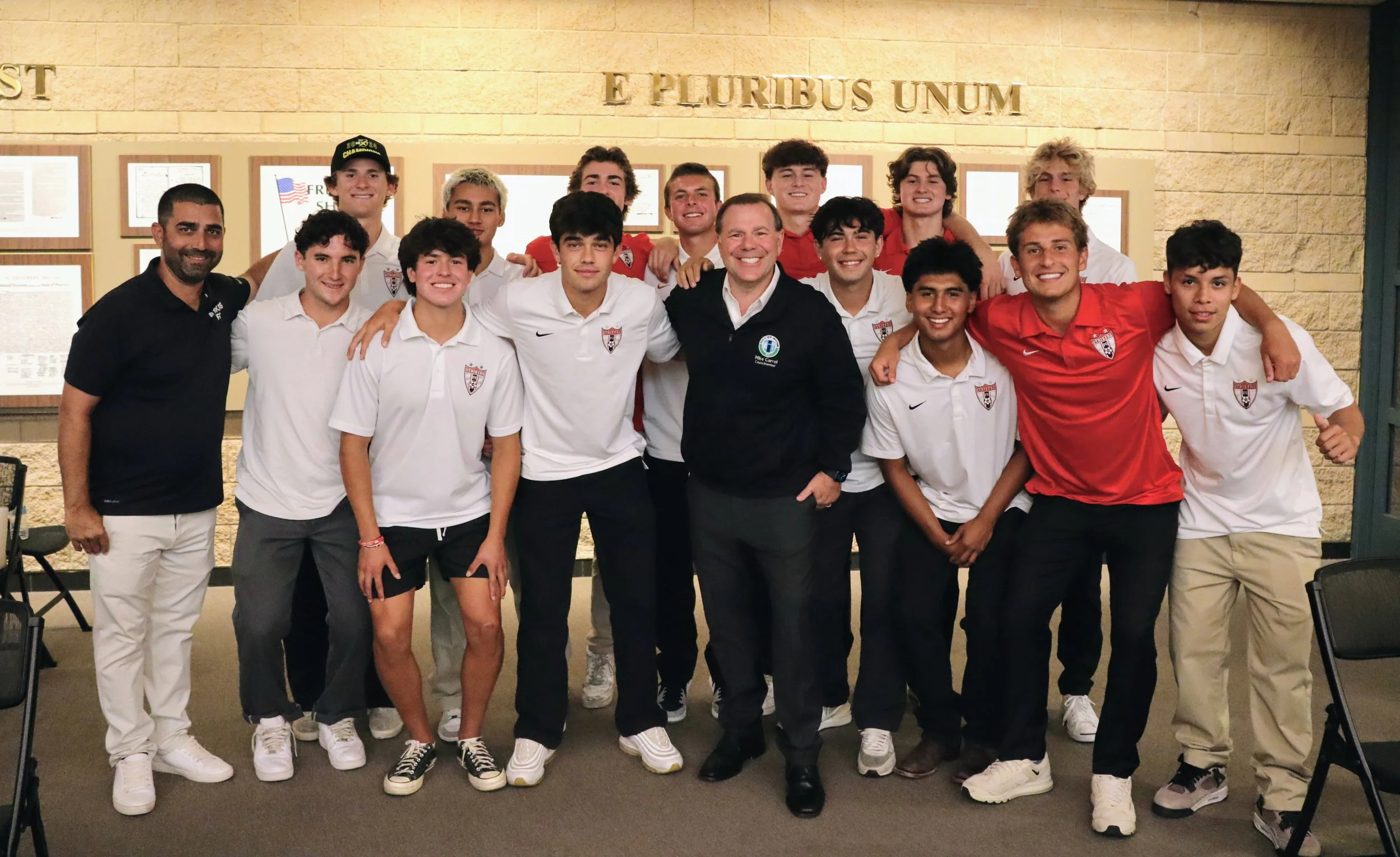 Group of young male athletes wearing white and red sports shirts, posing with two adults in a room with a brick wall and framed documents. The phrase 'E Pluribus Unum' is displayed on the wall.