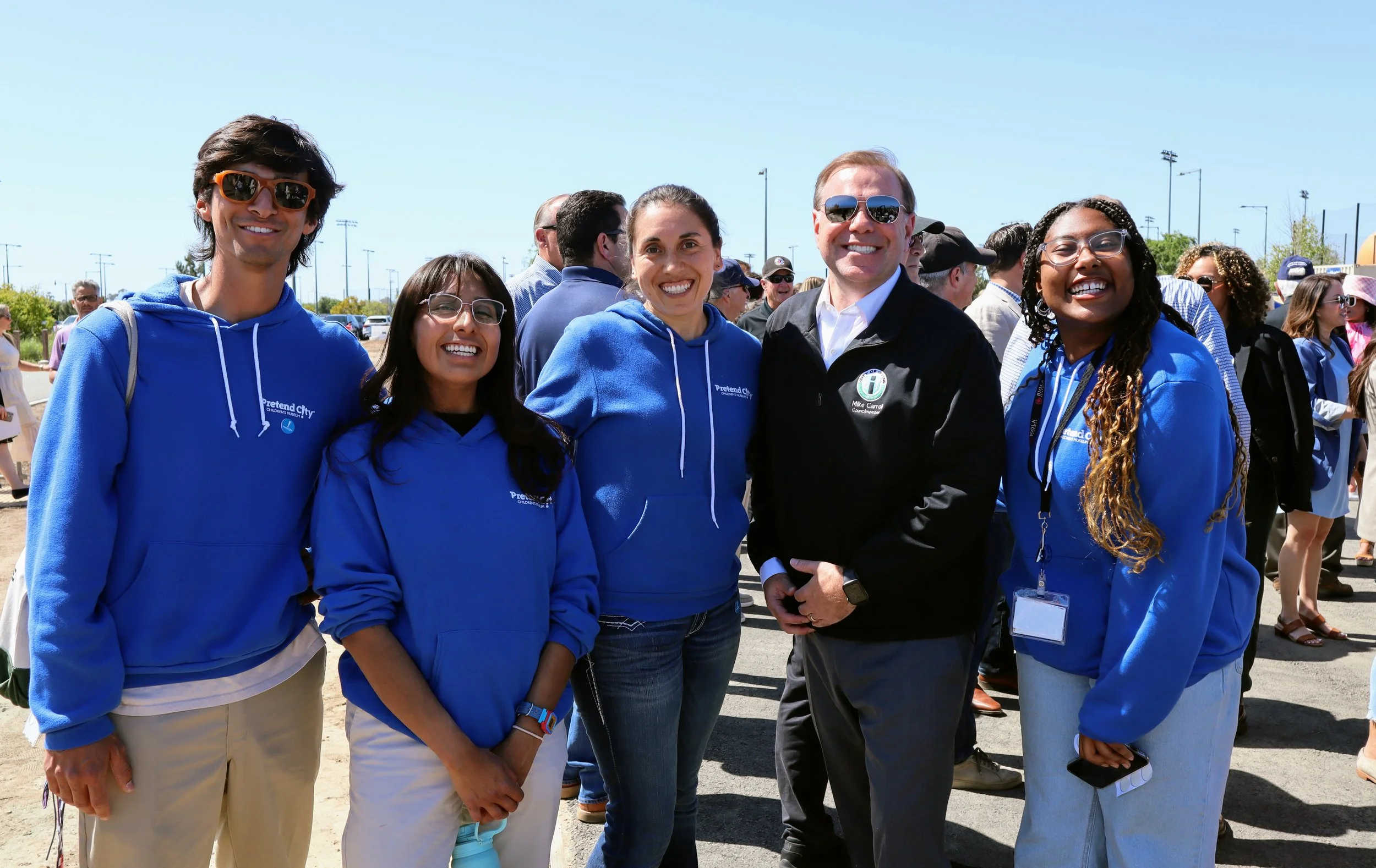 Group of five smiling people standing outdoors on a sunny day, wearing blue hoodies with a city logo, with other people in the background.
