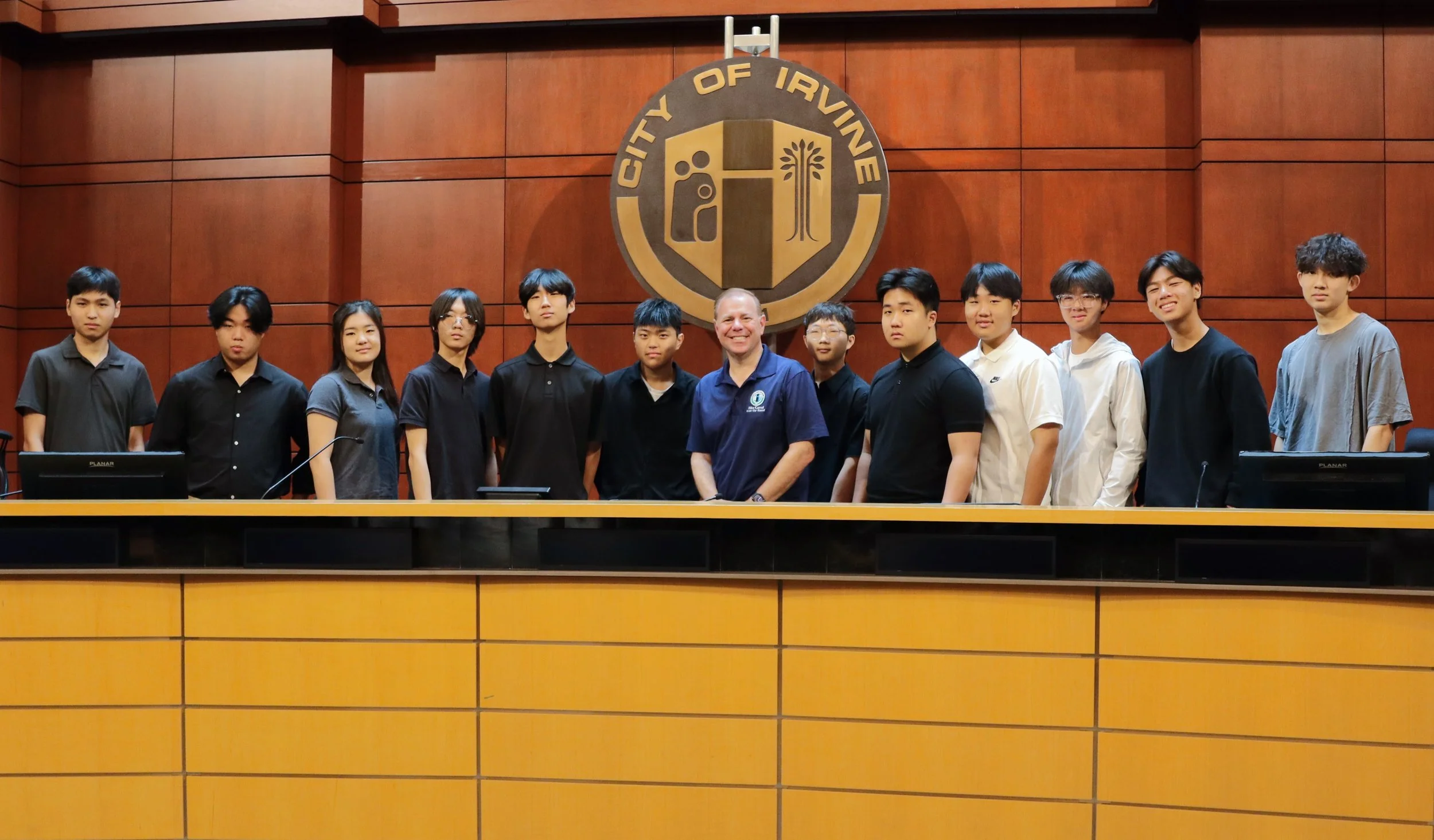 Group of students and a teacher standing behind a wooden courtroom bench in front of a wooden wall with a city emblem that reads "City of Irvine".