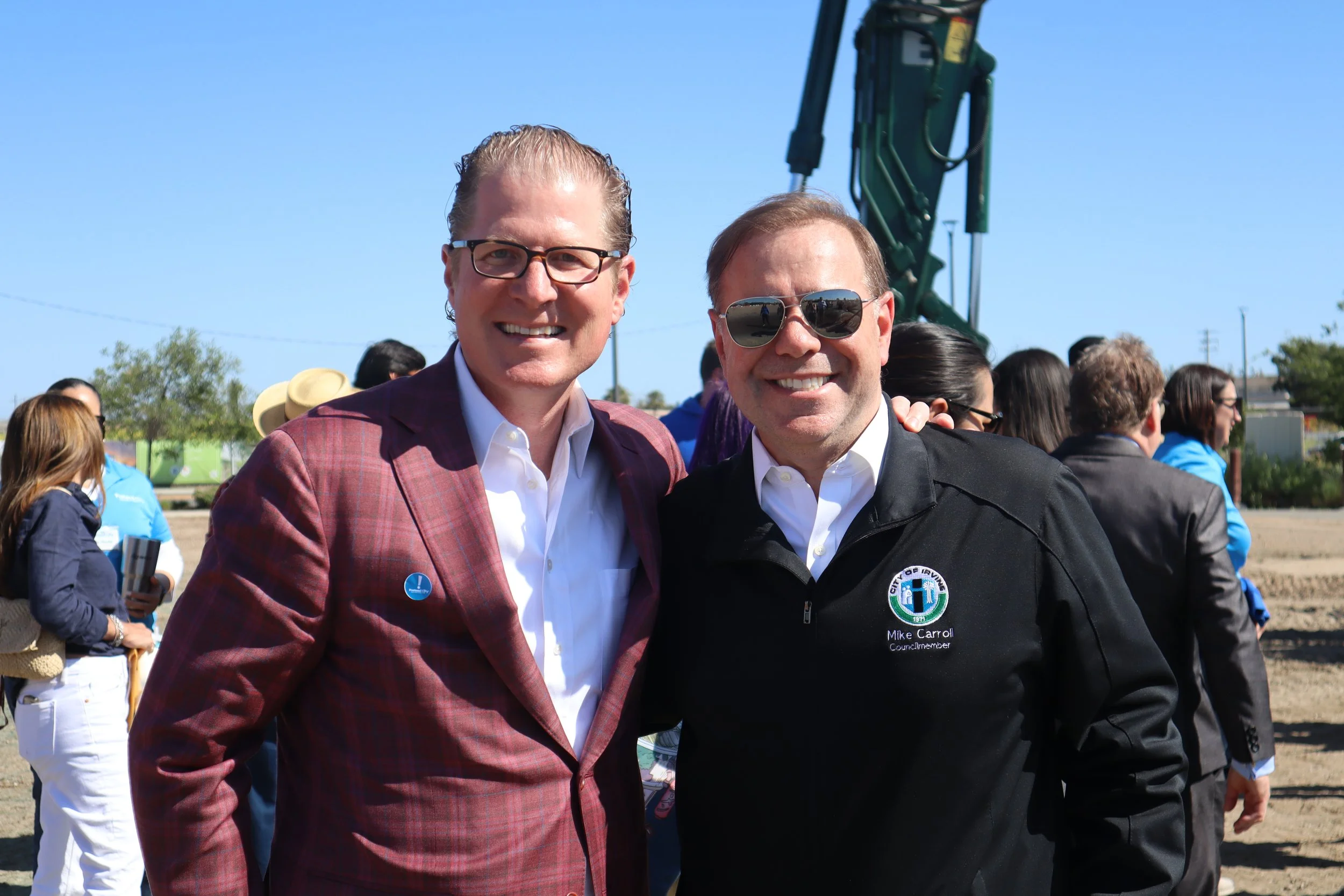 Two men smiling at the camera, one in a checked blazer and the other in a black jacket with sunglasses, at an outdoor event with a crowd and construction equipment in the background.