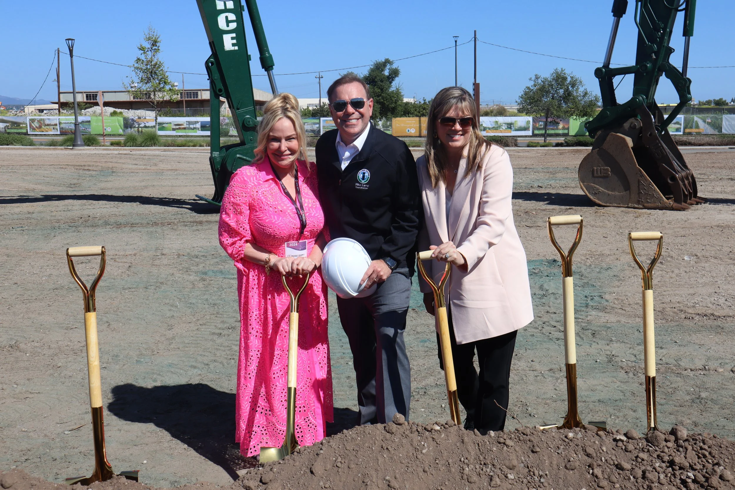 Three people standing at a groundbreaking ceremony with shovels and a pile of dirt, a large excavator in the background, on a construction site under a clear blue sky.