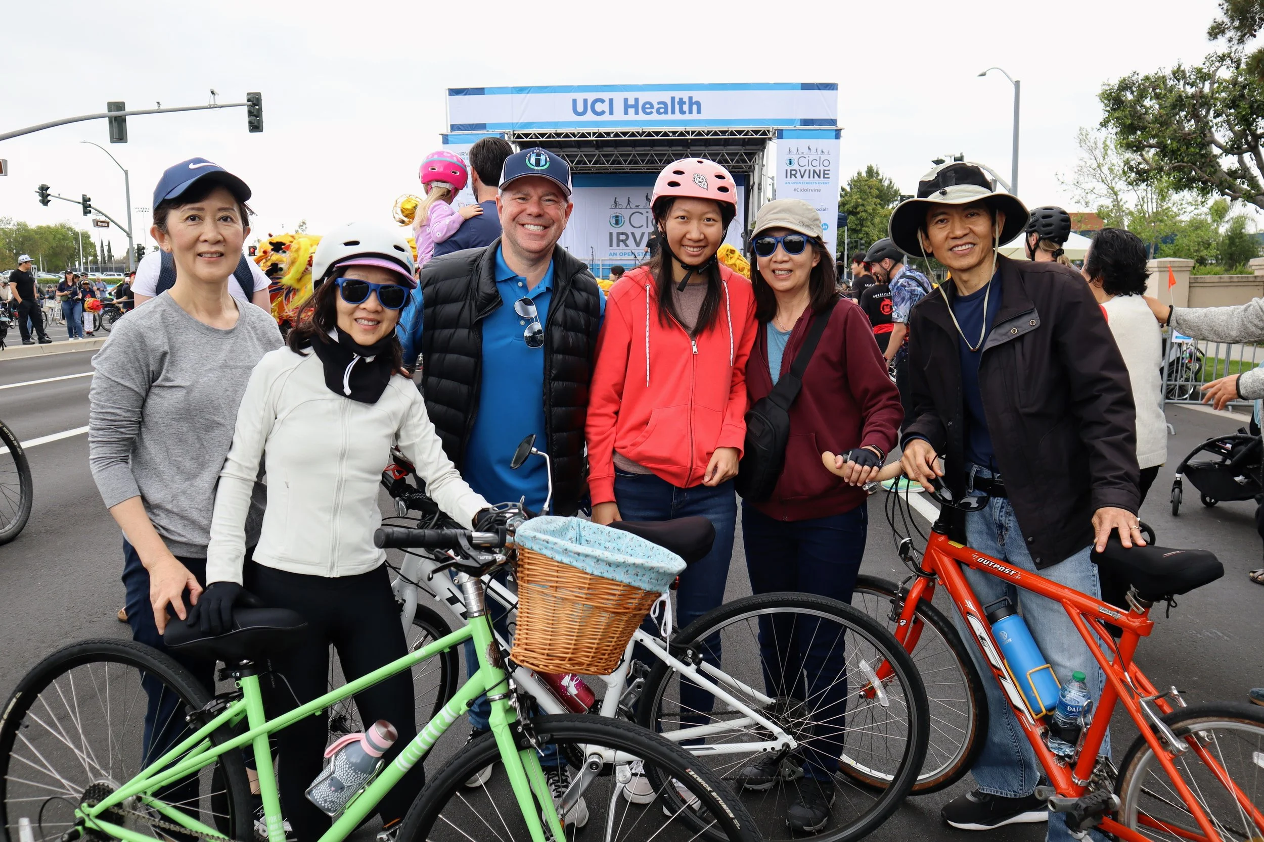 Group of six diverse adults standing with bicycles in front of a stage with a sign that reads 'UCI Health' at an outdoor event.