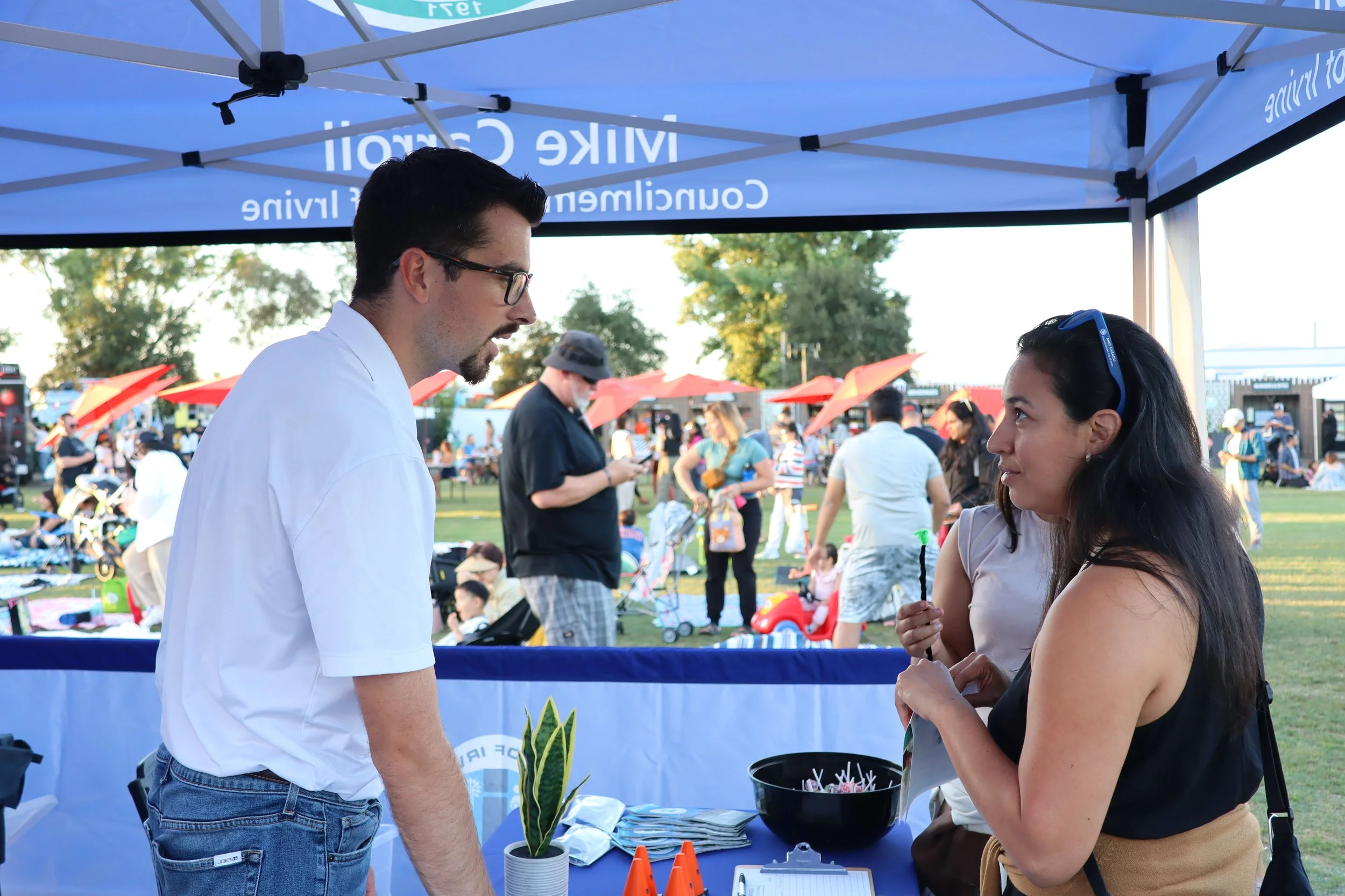 A man and woman talking at an outdoor informational booth with a blue canopy, surrounded by people at a community event or fair.