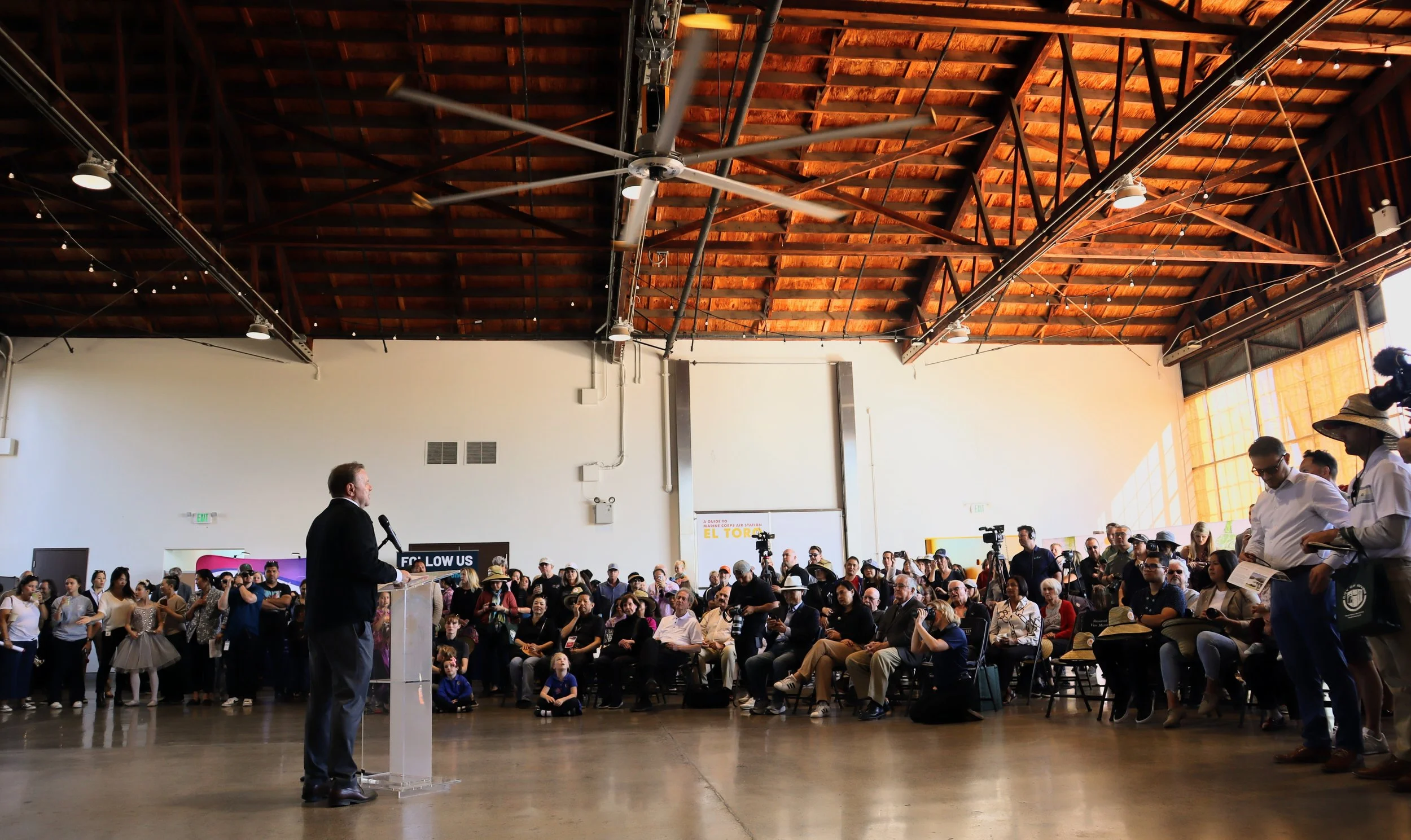A man giving a speech at a podium in a large indoor space with an audience seated in chairs and some standing around, under a high wooden ceiling with exposed beams.