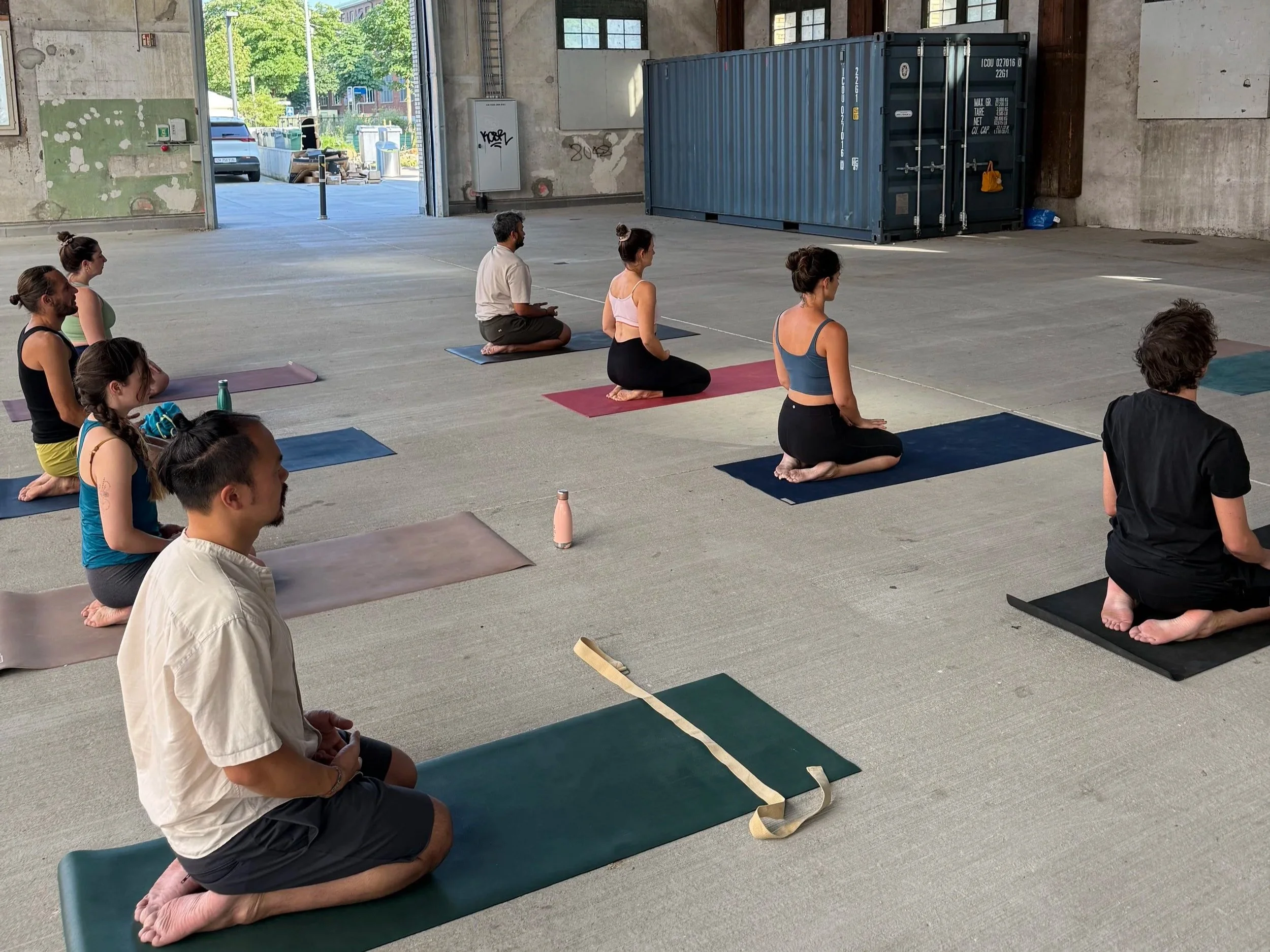 People participating in a yoga class inside a spacious, industrial-style warehouse with high ceilings, exposed beams, and large windows, sitting on mats in a meditative posture.