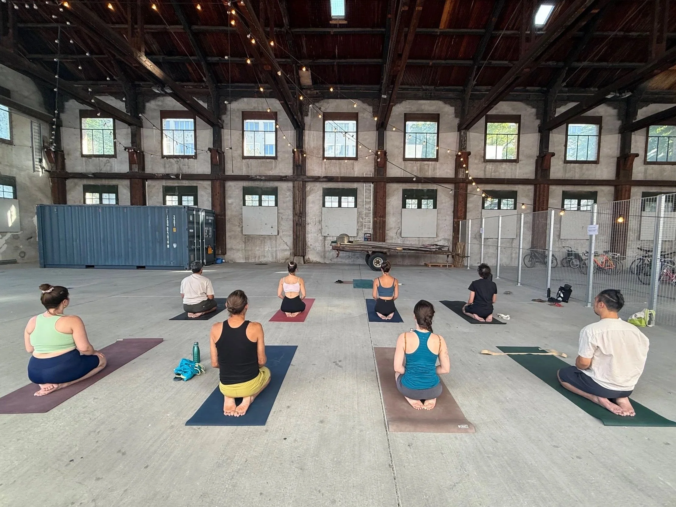 Group of people participating in a yoga class in an industrial-style warehouse with high ceilings, large windows, and string lights, viewed from behind.