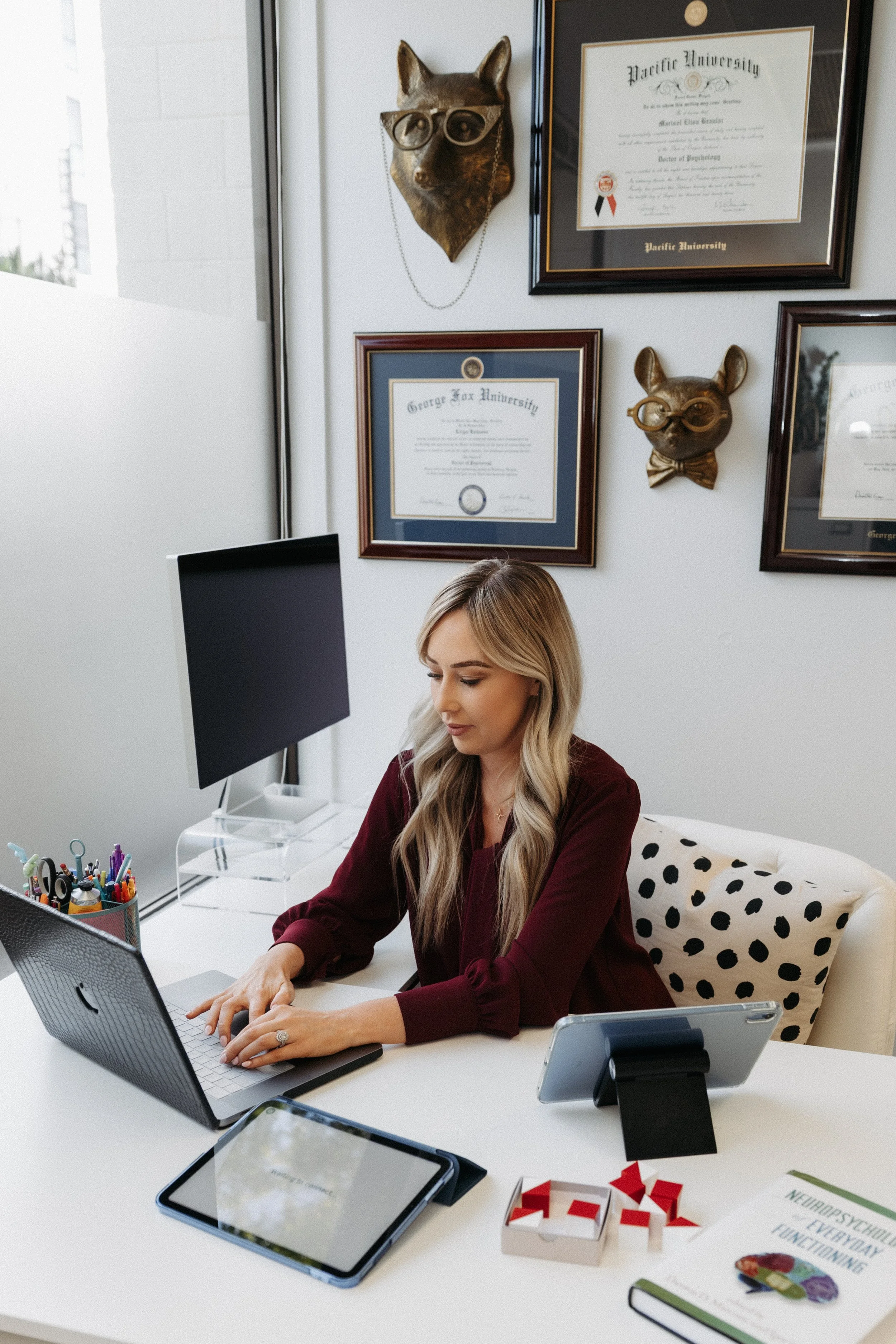 A woman with long blonde hair wearing a maroon blouse sitting at a white desk, working on a computer with a black monitor. The desk has a smartphone on a stand, a tablet, a container with colorful pens, and a game with red and white pieces. The background shows framed diplomas and a sculptural wall art of a fox head wearing glasses, along with framed certificates on the wall.