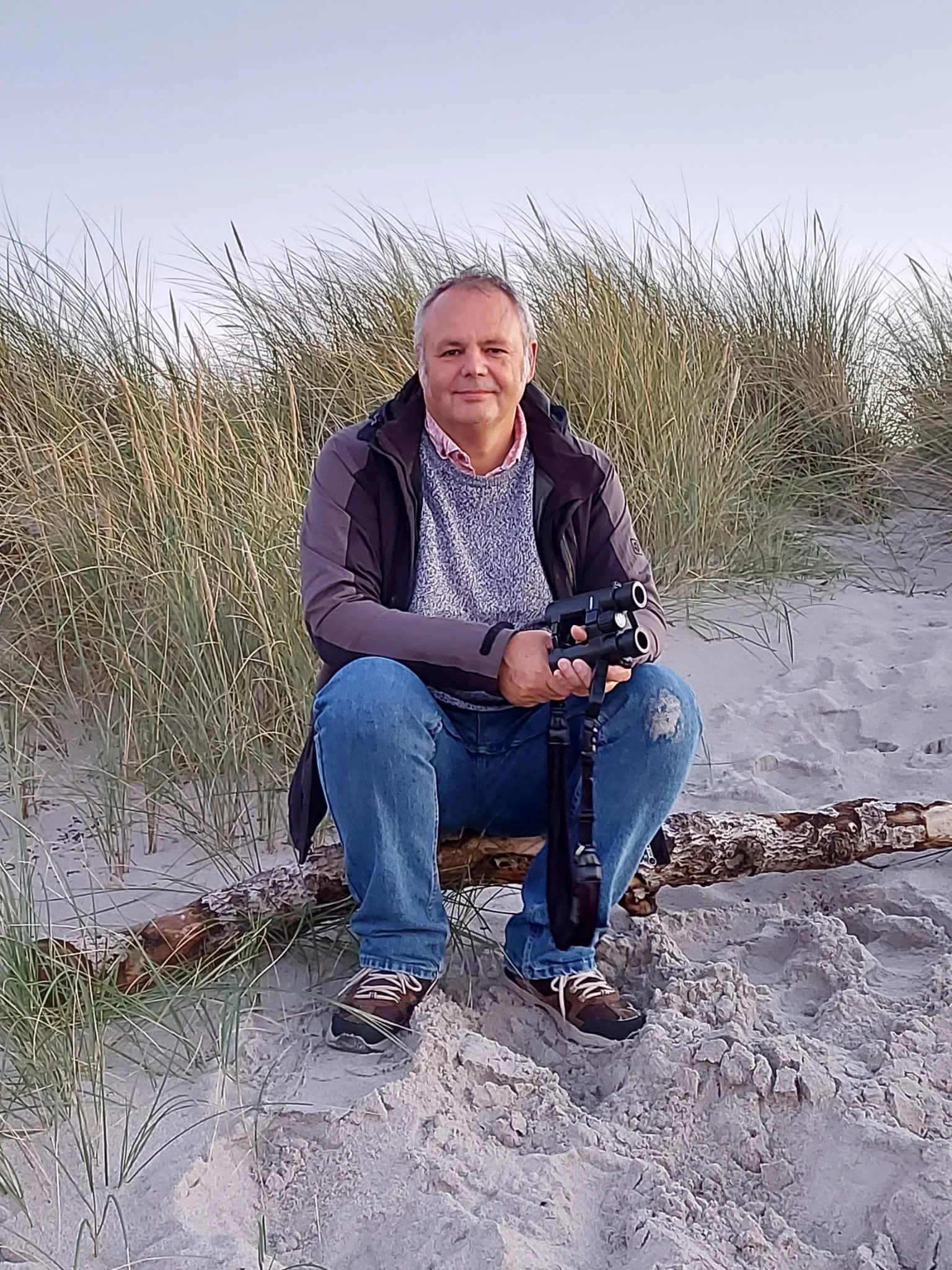 Professor Jeff Ollerton sitting among coastal sand dunes, holding a camera with a telephoto lens.