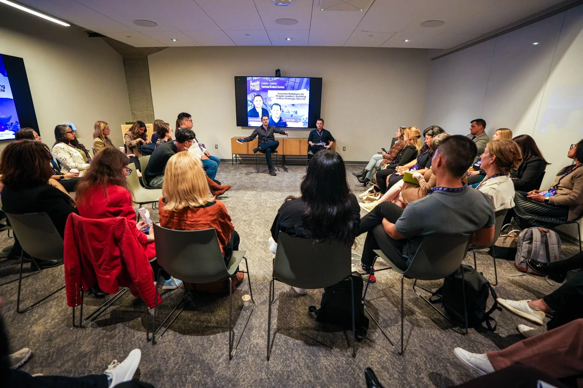 People attending a panel discussion in a conference room. There are two speakers seated at the front, with a large screen behind them displaying the event details. Attendees are seated in rows, some taking notes or looking at their phones.