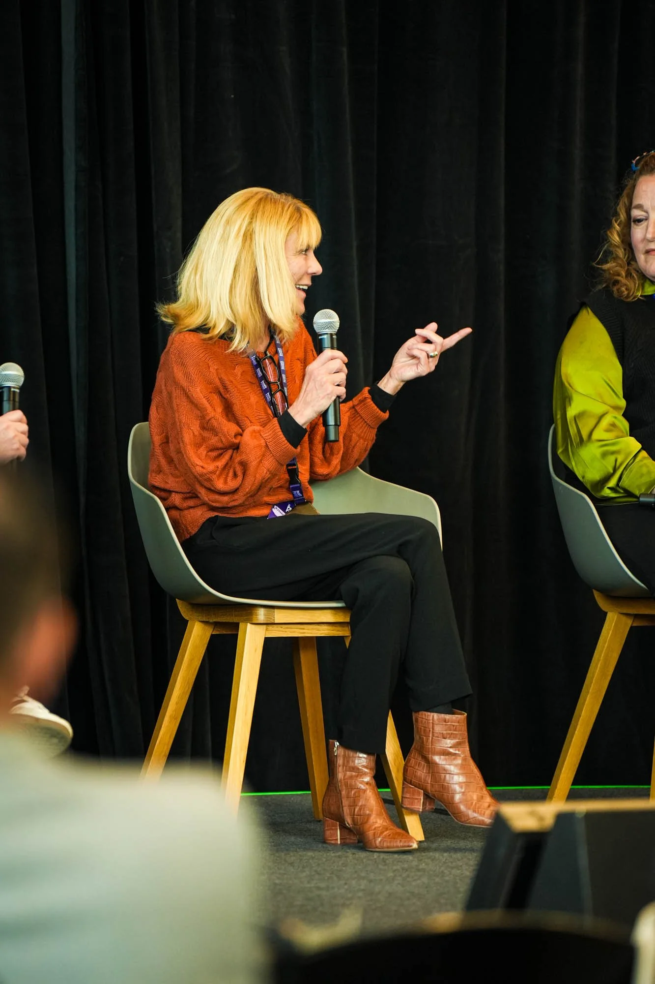 Women sitting on chairs participating in a panel discussion or interview, with one woman at the center holding a microphone, wearing a rust-colored sweater and brown boots, dark curtains in the background.