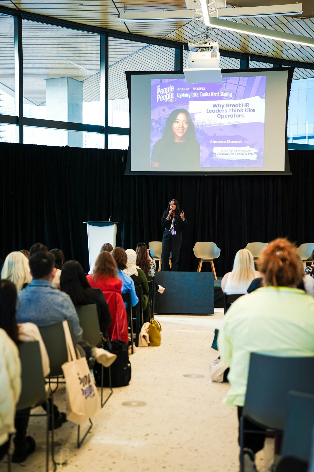 A woman is speaking on stage at a conference in front of a large screen showing a presentation slide titled "Why Great HR Leaders Think Like Operators". The audience is seated facing the stage in a modern, glass-walled conference room with a high cei