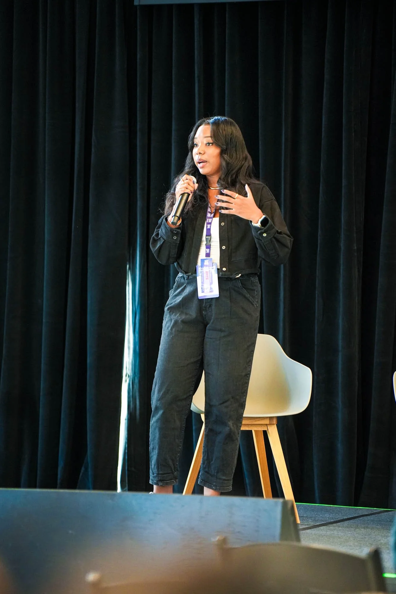 A woman with black, wavy hair speaking into a microphone on stage in front of black curtains, wearing a black jacket, white shirt, and black pants, with a purple lanyard and a smartwatch.