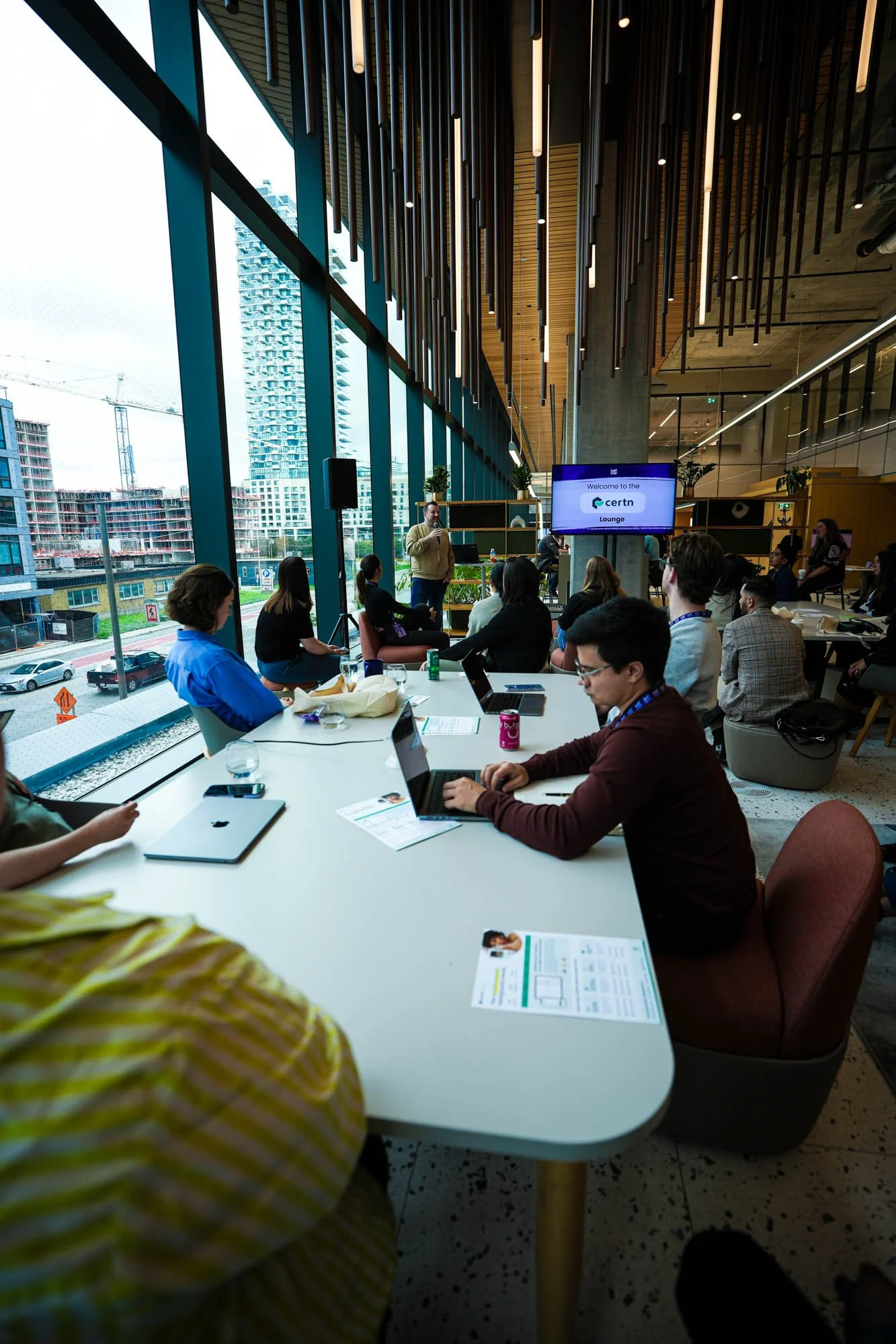 People participating in a meeting inside a modern conference room with large floor-to-ceiling windows showing a cityscape, some are using laptops and papers, and a speaker is presenting in front of a screen that says 'Welcome to the Certn Lounge'.
