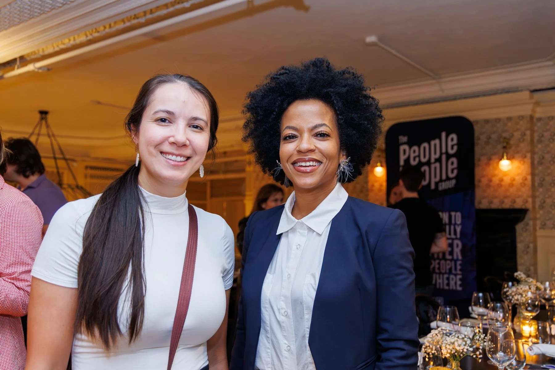 Two women smiling at an indoor event, one with long dark hair in a white top and the other with curly black hair wearing a navy blazer and white shirt, with event signage and tables in the background.