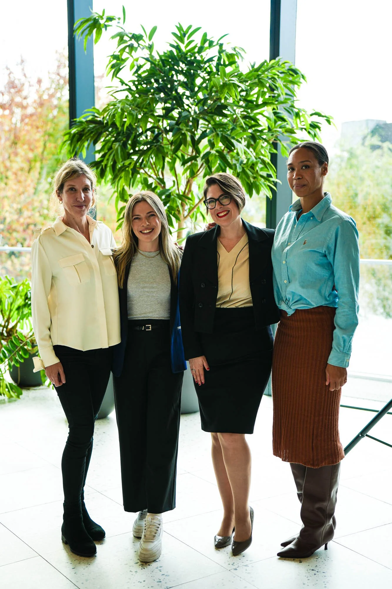 Four women standing indoors near large windows with green plants and trees outside, smiling and posing for a photo.