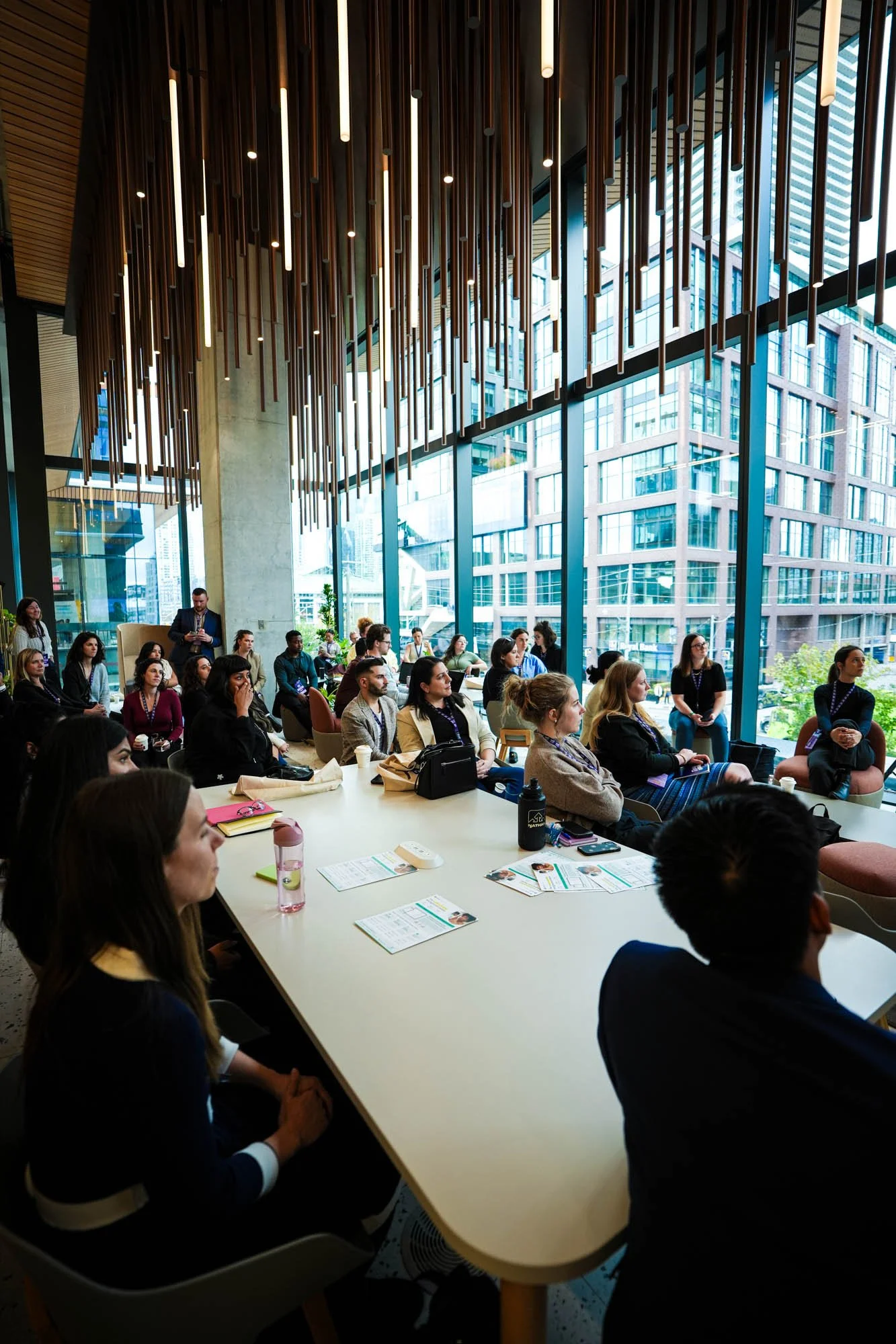 A group of diverse people attending a conference or seminar in a modern building with large glass windows and city view, sitting at a long white table with papers, laptops, and water bottles.