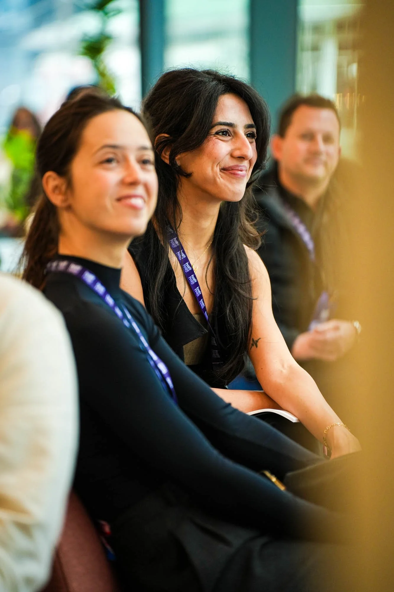 Three people sitting at a table, smiling, with large windows and green plants in the background.