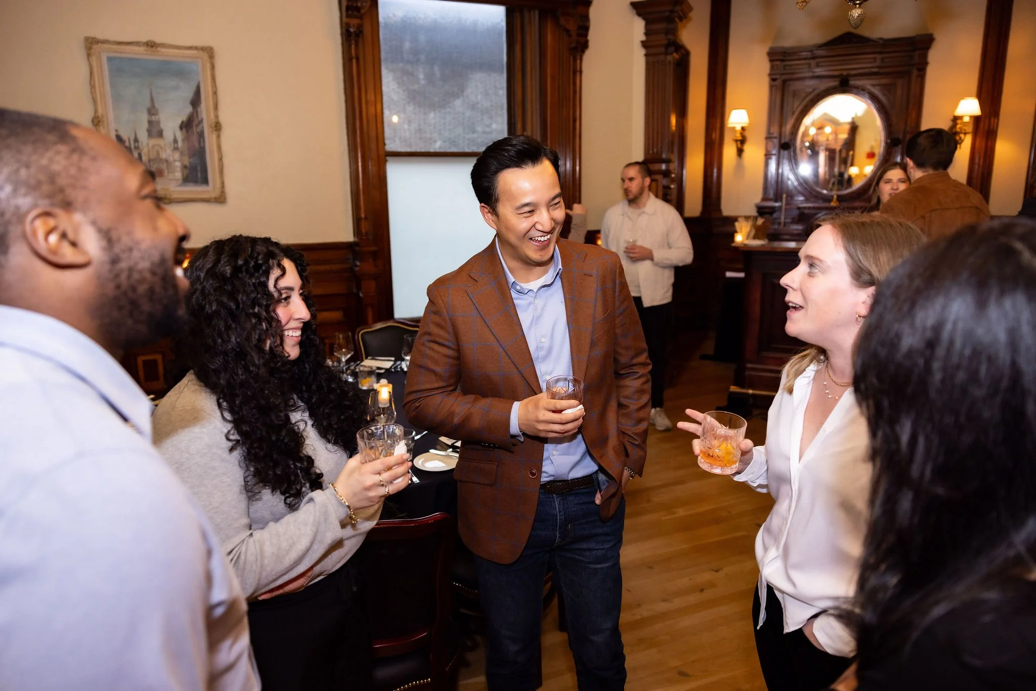 People socializing at a gathering or party in a warmly lit room with wood trim. They are holding drinks and engaging in conversation, with a man in a checkered blazer smiling at a woman.