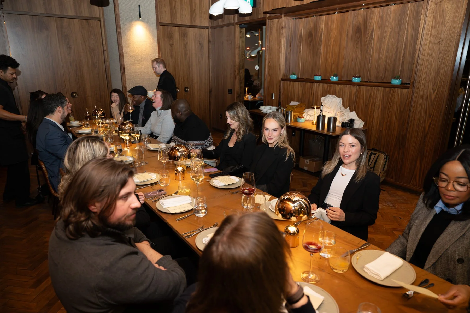 A group of people sitting at a long dining table in a warmly lit wooden-paneled restaurant, engaging in conversation and enjoying drinks and meals.