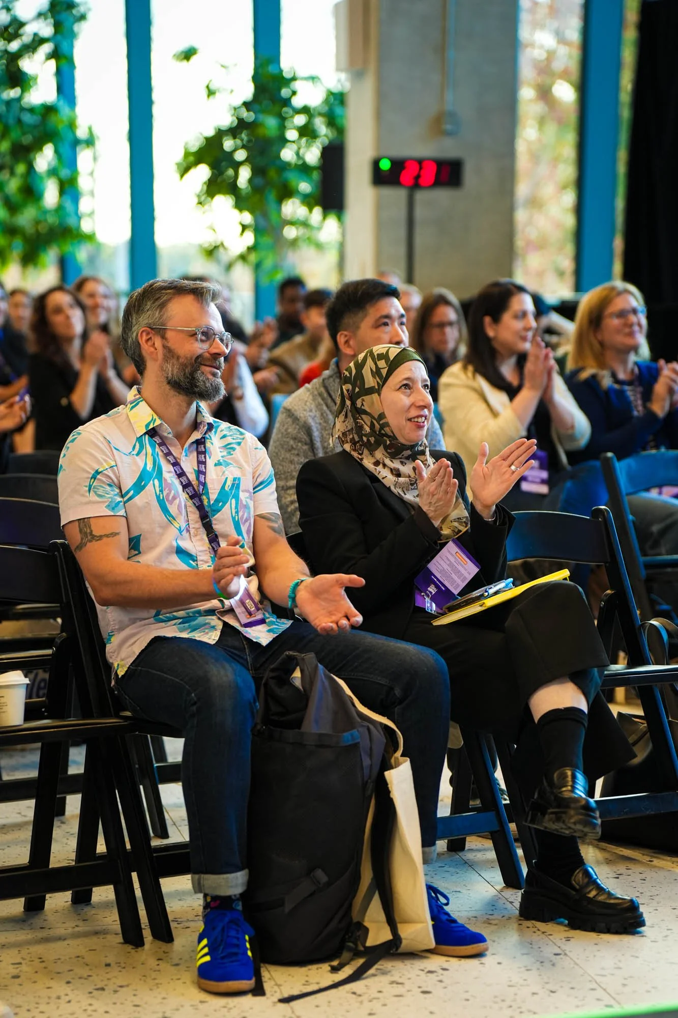 Audience attending a conference, sitting and clapping in a room with large windows and greenery outside, with a digital timer showing 2:11 in the background.
