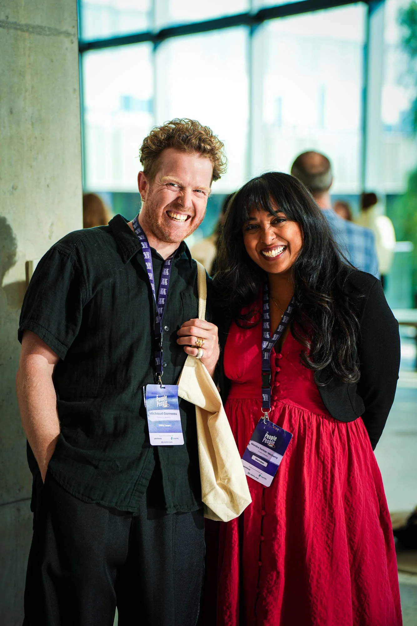 Two smiling conference attendees, a man with curly hair and a woman with long black hair, posing together at a conference called 'People People,' both wearing conference badges around their necks. The man is dressed in a black shirt and the woman in 