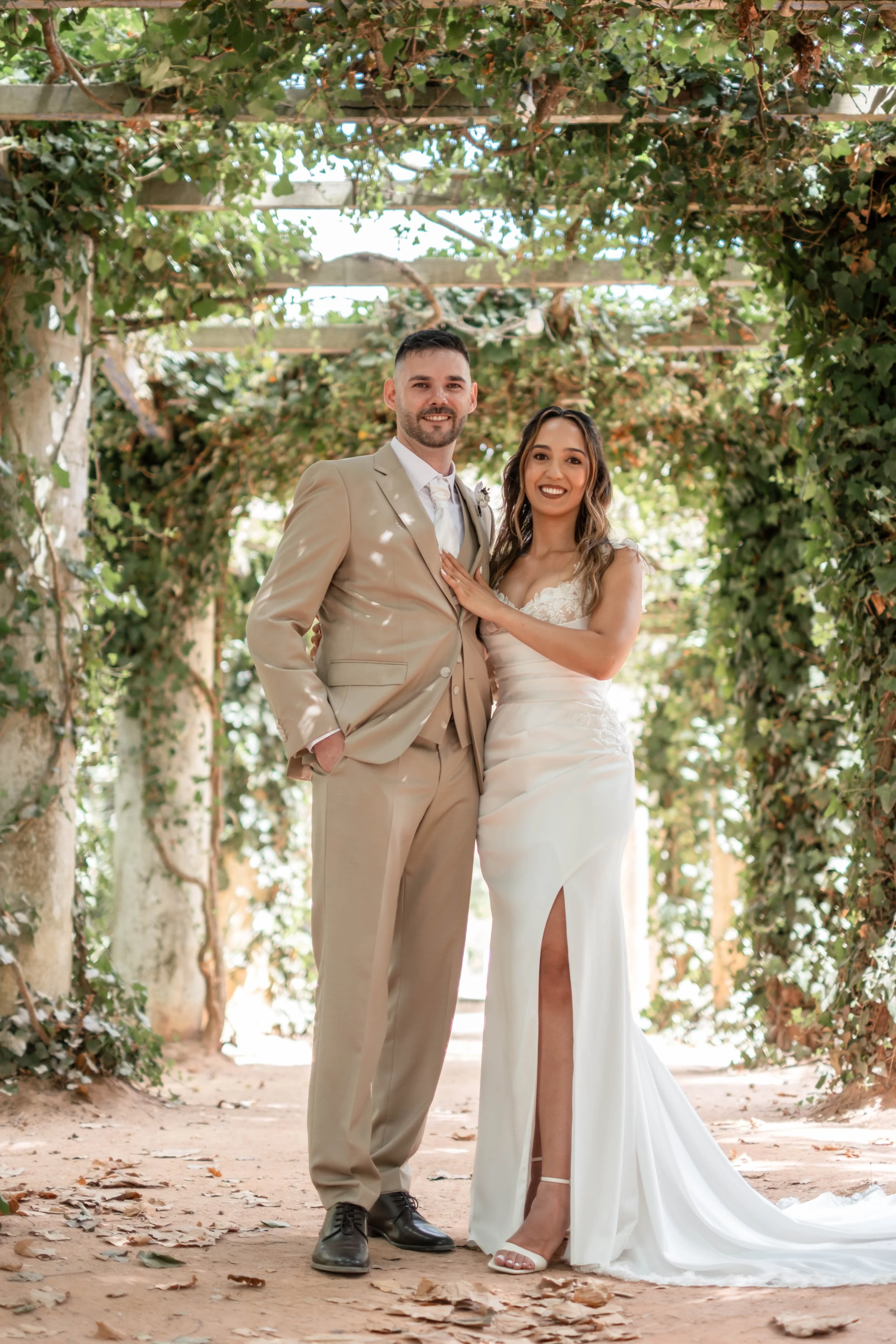 Noivos em retrato de corpo inteiro sob uma pérgola coberta de vegetação